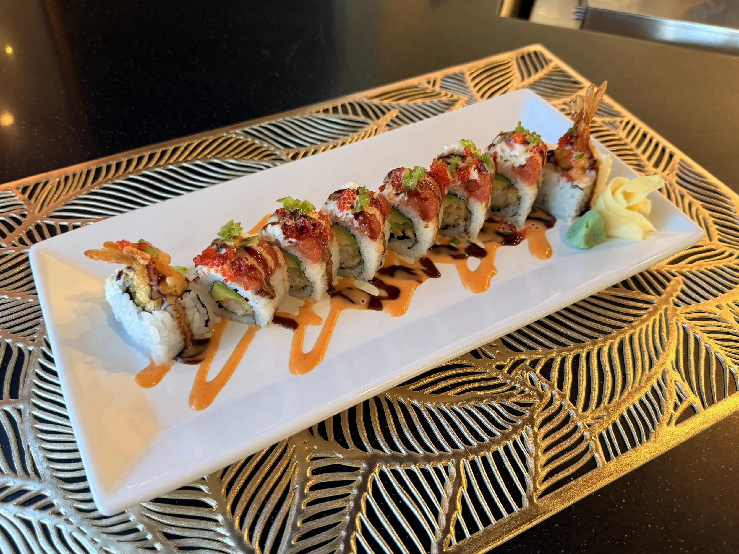 A plate of sushi rolls garnished with green onions, drizzled with sauce, and served with wasabi and pickled ginger on a decorative tray.