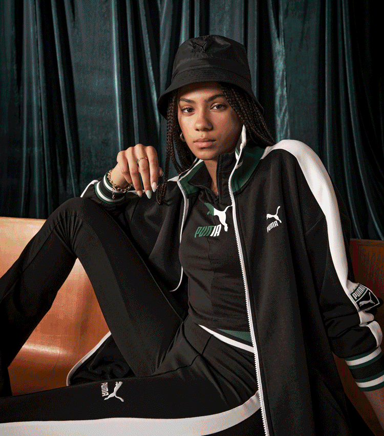 A young woman wearing a black and white Puma tracksuit, black bucket hat, and sitting on a bench with her leg crossed in front of a dark curtain background.
