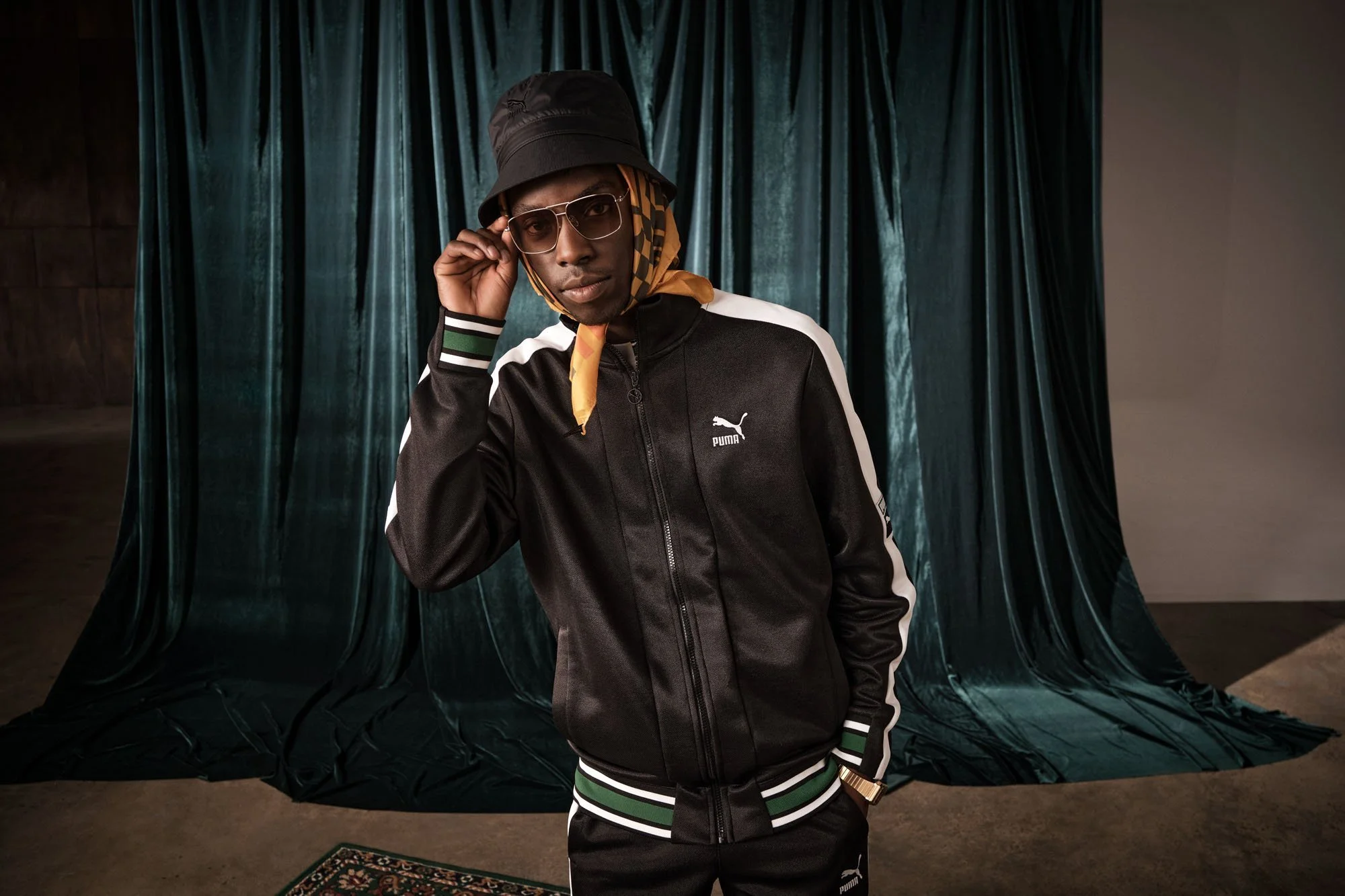 A young man wearing a black Puma jacket, black bucket hat, yellow scarf, glasses, and a gold watch posing in front of a dark green velvet curtain.