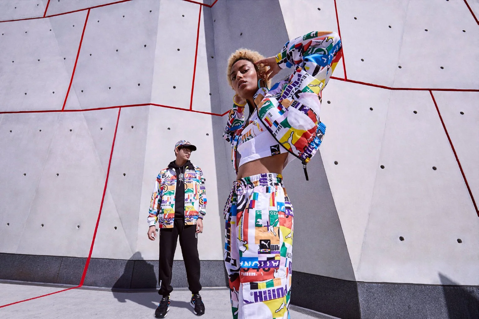 Two models wearing colorful, graphic print streetwear pose against a modern concrete wall with red geometric lines. One is a woman in a crop top and wide-leg pants, the other is a man in a jacket and pants, both sporting accessories and sneakers.