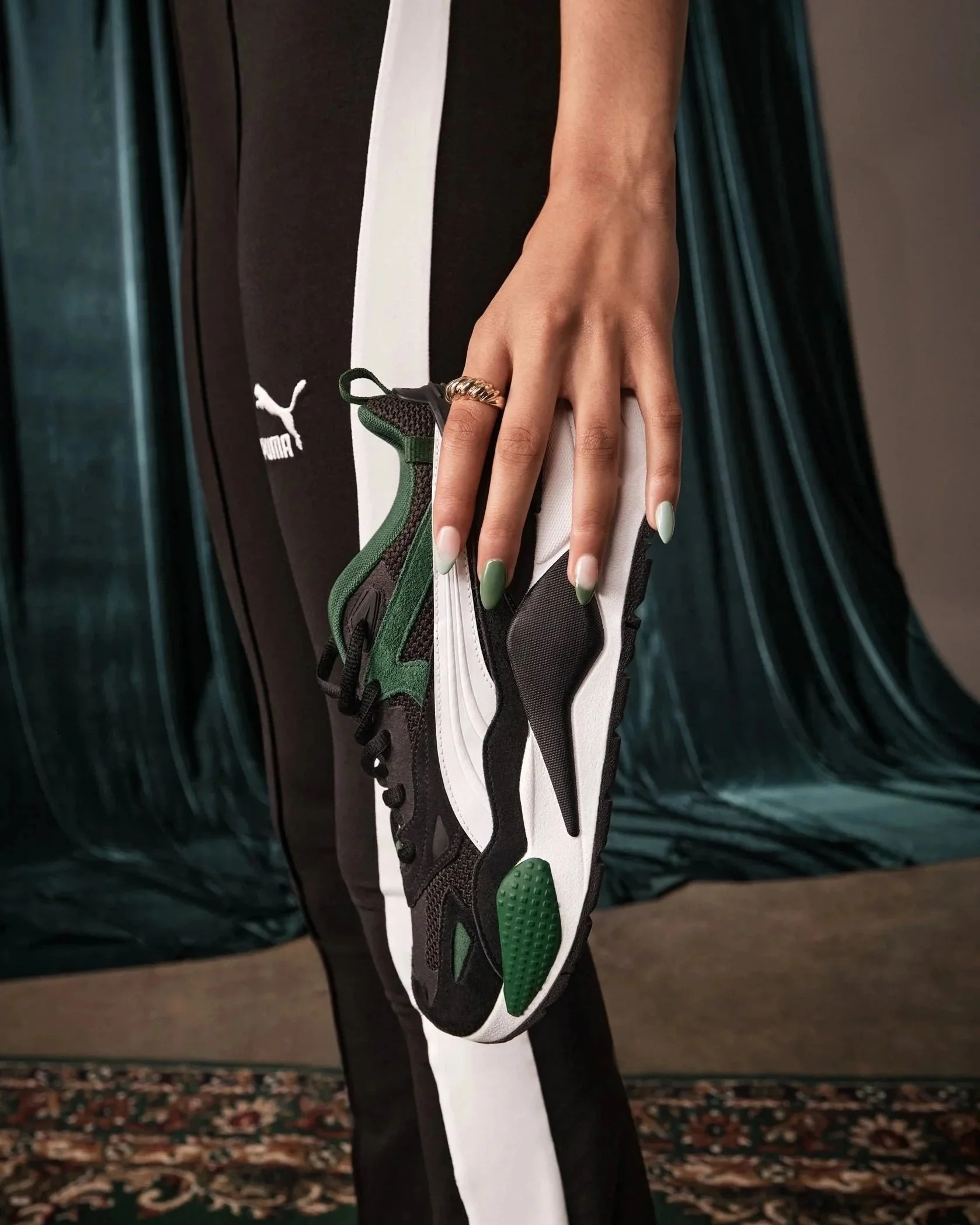 A person resting their hand on a black and white puma sneaker with green accents, wearing a gold ring and painted nails, with a dark curtain and patterned rug background.