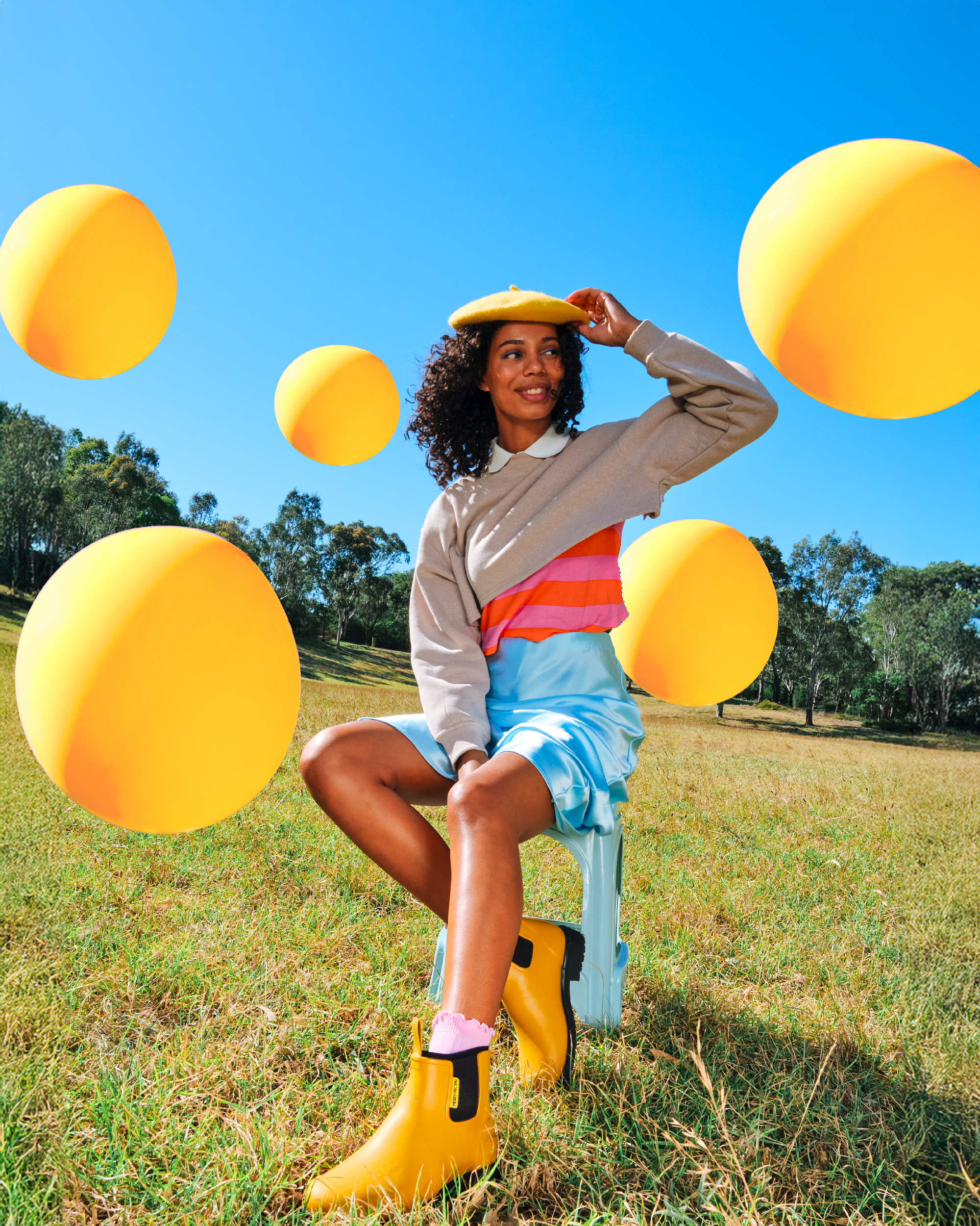 A woman sitting on a yellow ride-on toy in a grassy park, surrounded by large yellow plastic balls, on a sunny day with clear blue sky and trees in the background.
