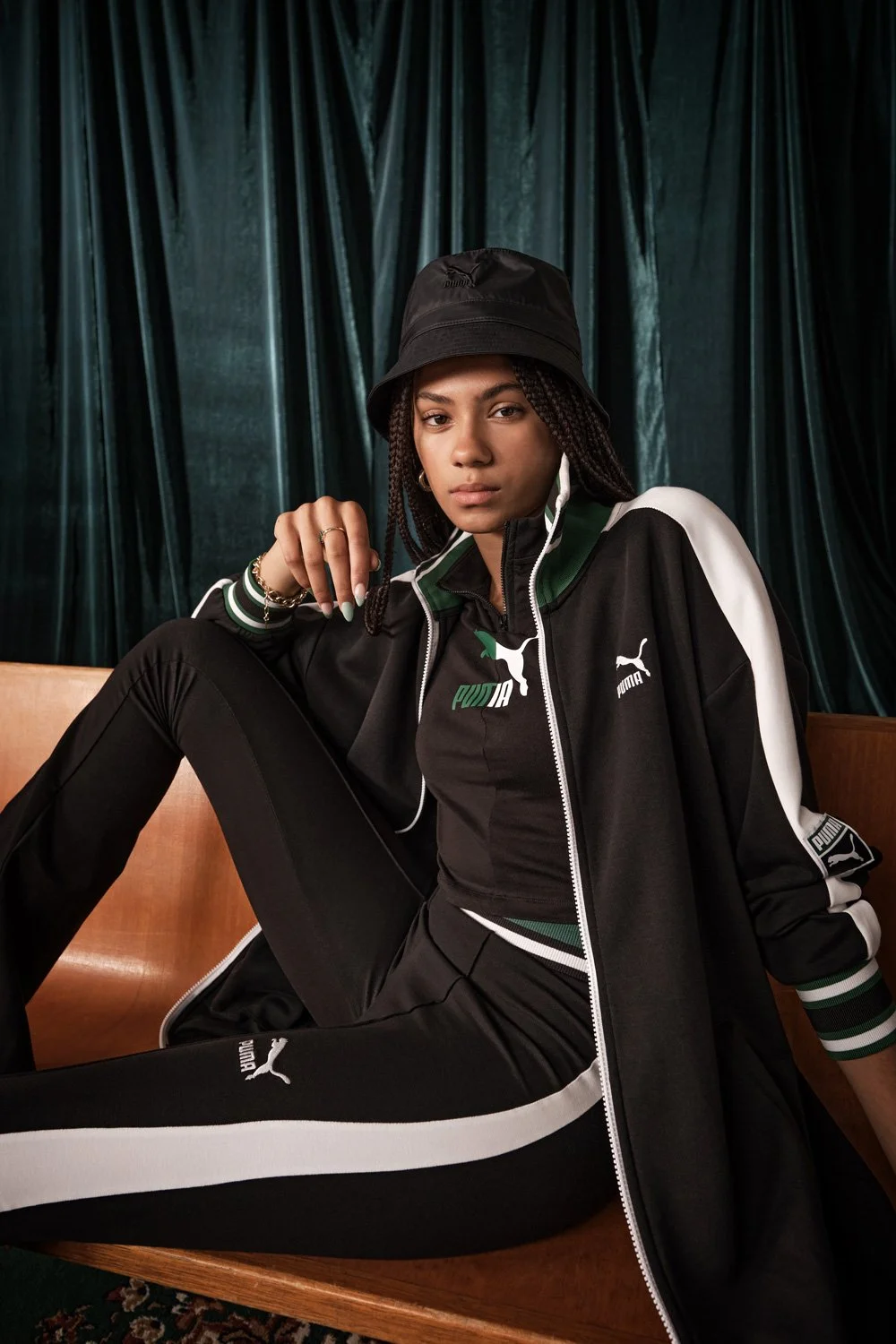 A young woman sitting on a wooden bench, wearing a black Puma tracksuit with green and white accents, a black bucket hat, and jewelry, in front of a dark green curtain.