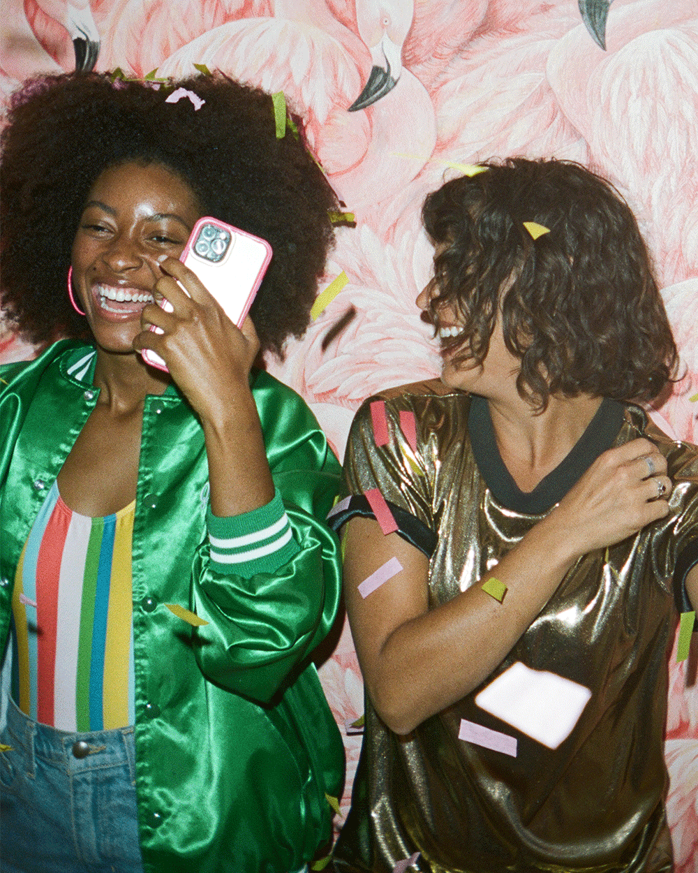 Two women smiling and having fun at a party. One woman with curly black hair, wearing a green satin jacket and a rainbow-striped top, is holding a smartphone to her ear. The other woman with shoulder-length curly dark hair, wearing a shiny gold shirt with pink and yellow paper confetti on her hair and sleeves, is laughing and looking at her friend. A pink flamingo patterned backdrop is behind them.