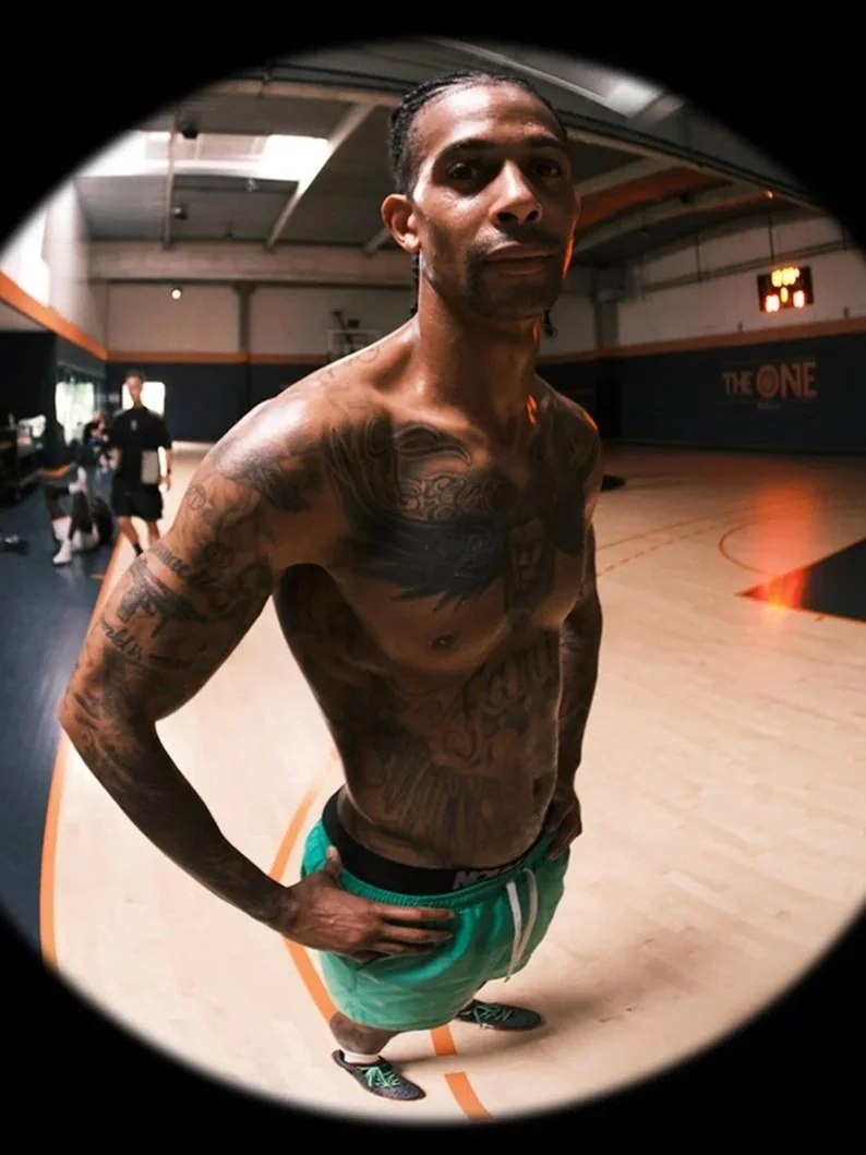 A shirtless man with tattoos on his chest, arms, and stomach stands on an indoor basketball court, looking into a fisheye lens camera. He is wearing green shorts and black athletic shoes. The background shows a gym environment with a scoreboard and a few other people.