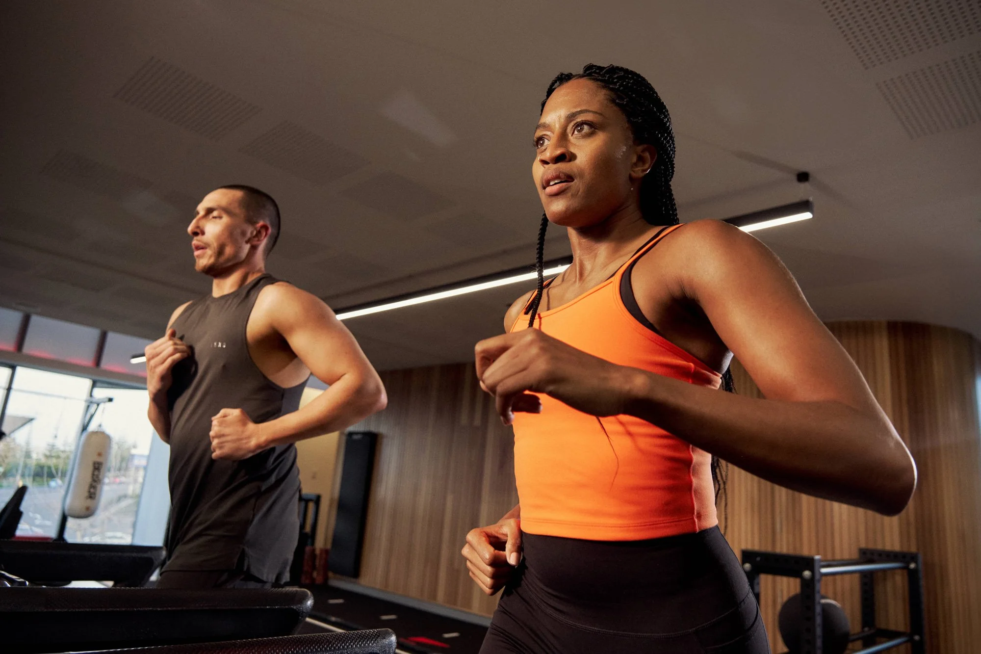 Two people running indoors, one woman in an orange sleeveless top and a man in a dark sleeveless shirt, both appearing focused.