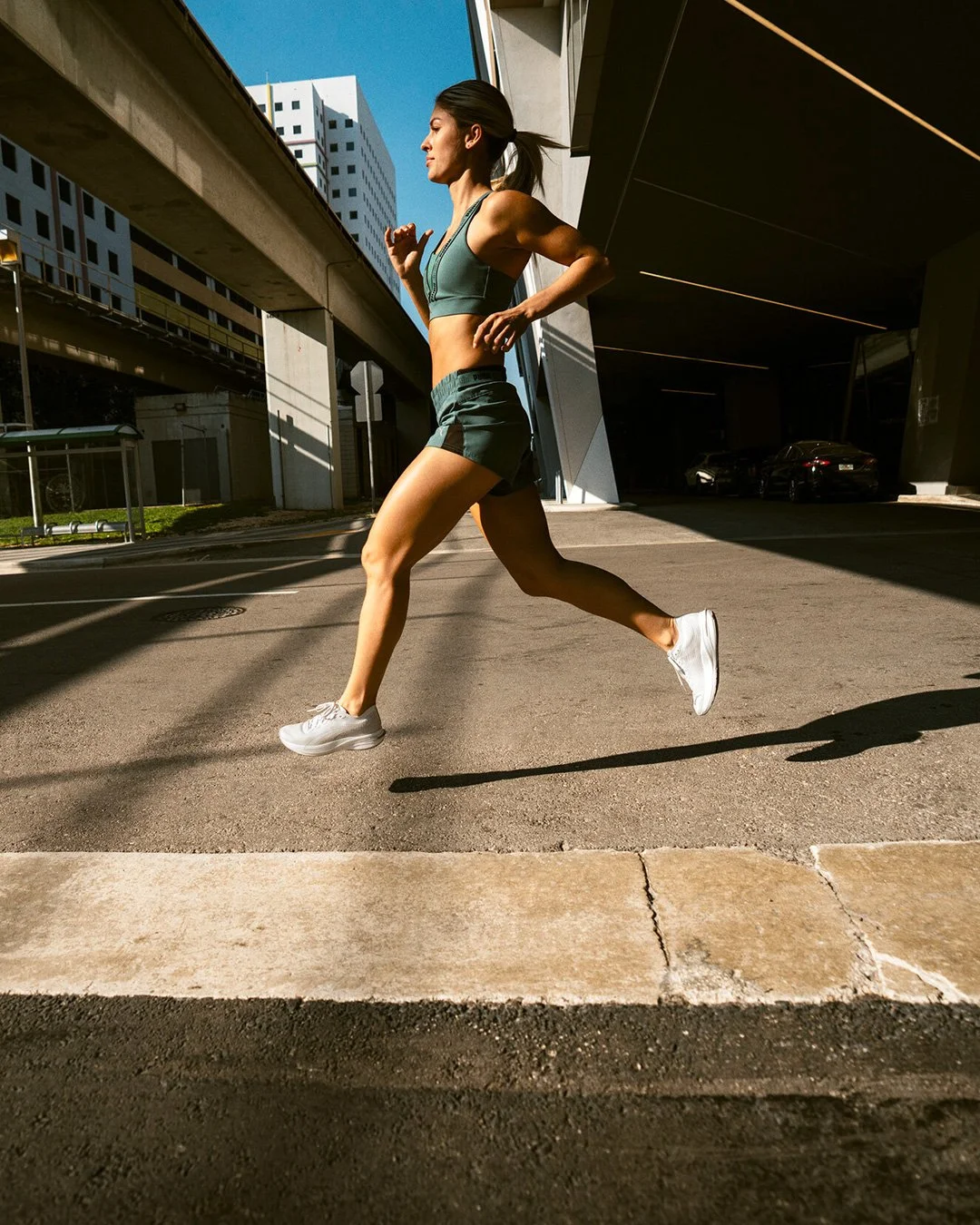 A woman running outdoors in athletic wear, shadow cast on the ground, under a bridge with modern buildings in the background.