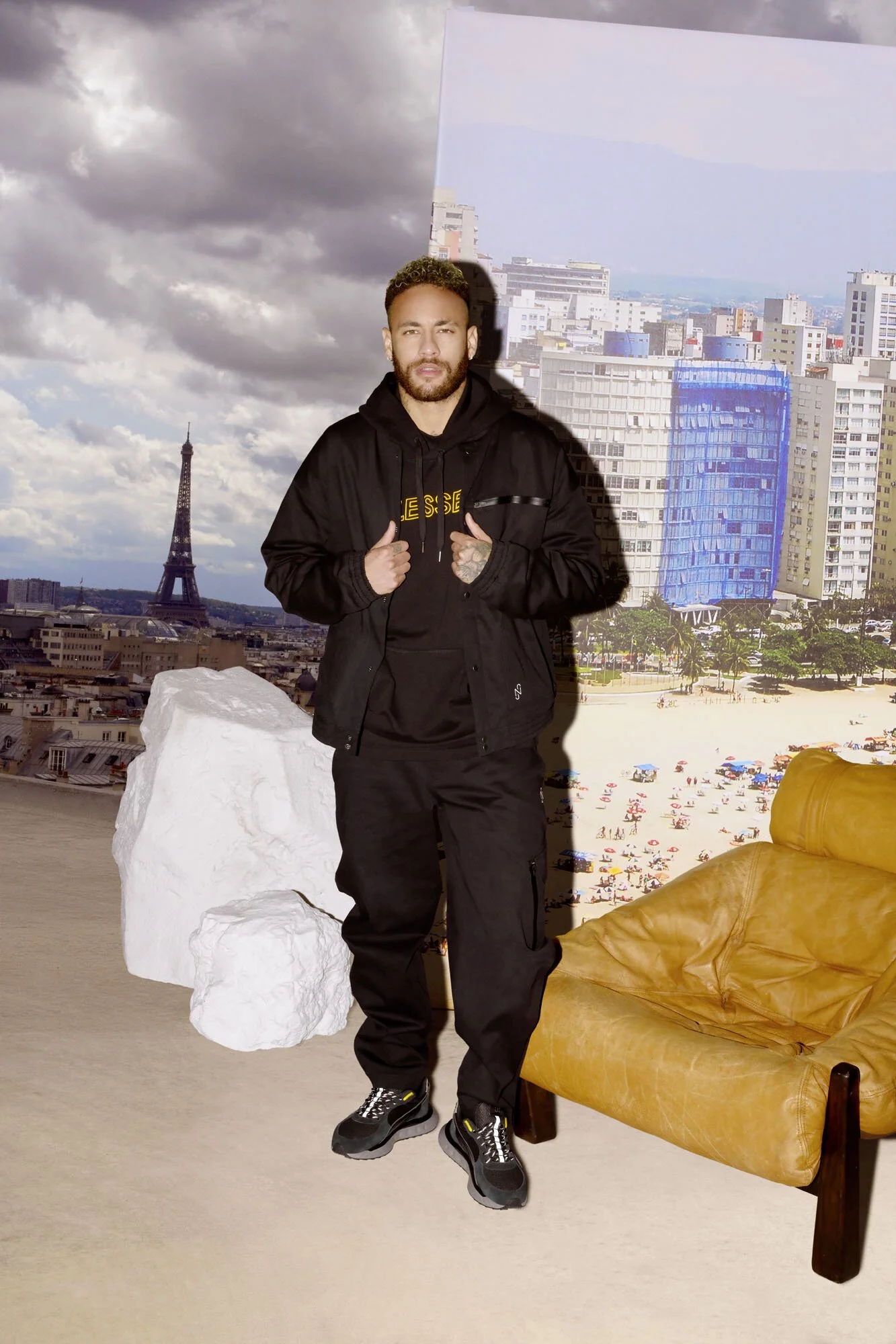 Neymar Jr wearing a black hoodie and pants, standing indoors with a background of a cityscape featuring the Eiffel Tower, clouds, and a sandy beach. There is a large white rock and a brown leather armchair in the foreground.