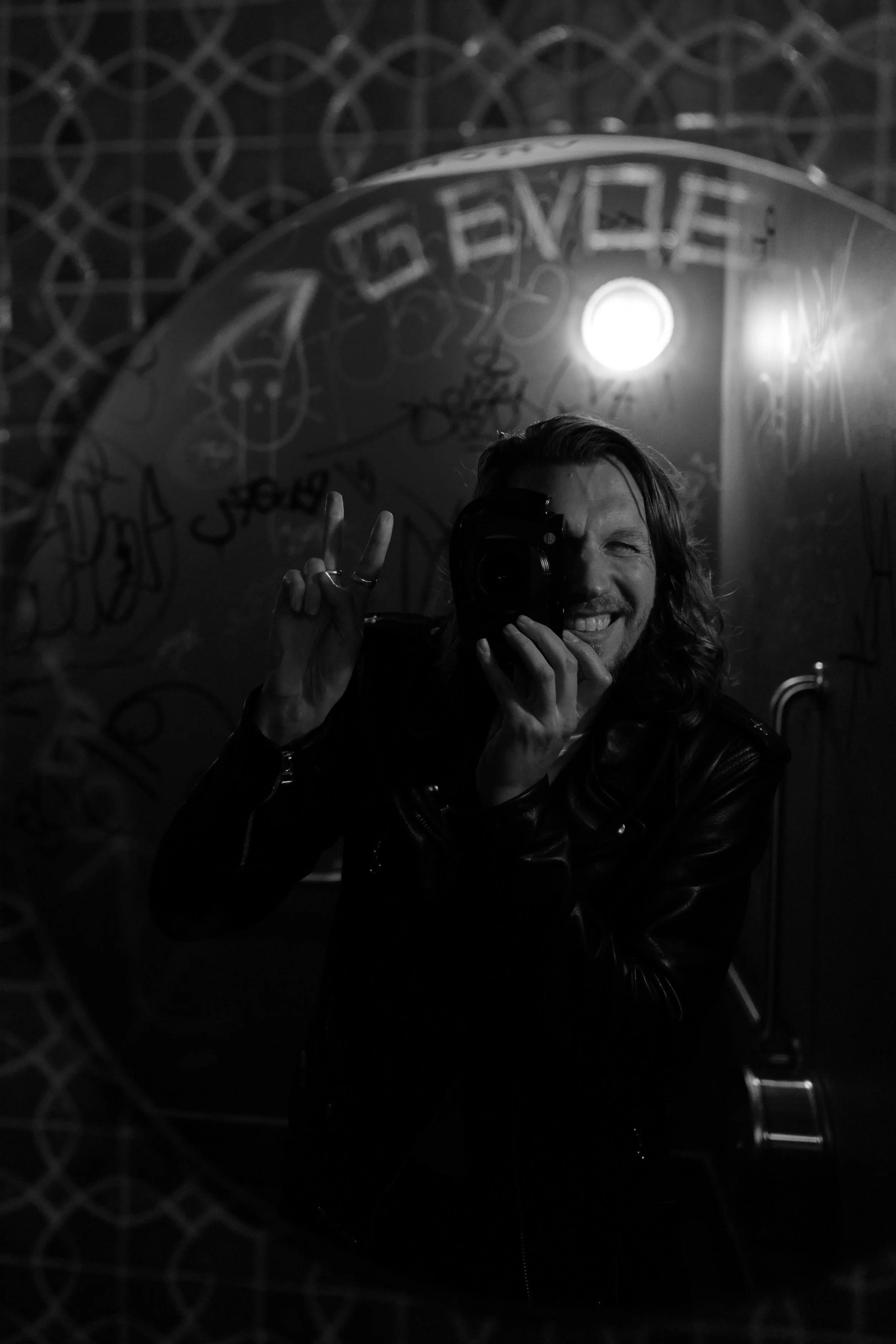Black and white photo of a smiling man with long hair taking a selfie in a mirror. He is holding a camera with one hand and making a peace sign with the other hand. The background includes graffiti on the wall and a bright light.