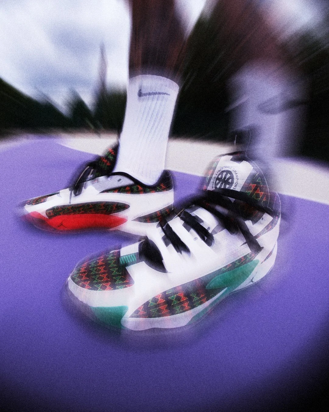 Pair of Nike sneakers with a black, red, green, and white pattern on a purple surface, with a white sock featuring a Nike logo and a basketball with a logo in the background.