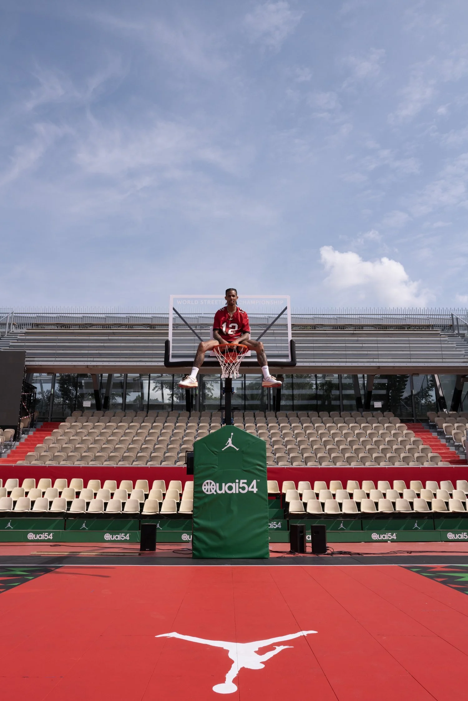 An empty basketball court with a red playing surface featuring a white Jumpman logo at the center. In the background, a person wearing a red sports uniform is sitting on a basketball hoop's rim, with a large backboard and a green padding labeled 'quai54.' Empty stadium seats are visible behind the court, along with a clear sky with some clouds.