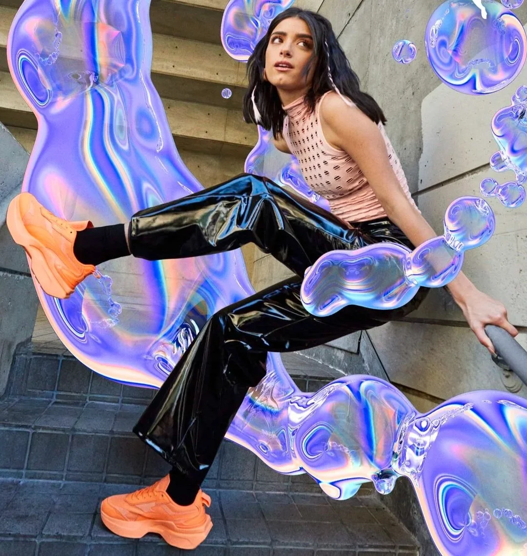A young woman in a pink, sleeveless top and shiny black pants with orange sneakers posing against a concrete wall with iridescent, swirling digital effects surrounding her.