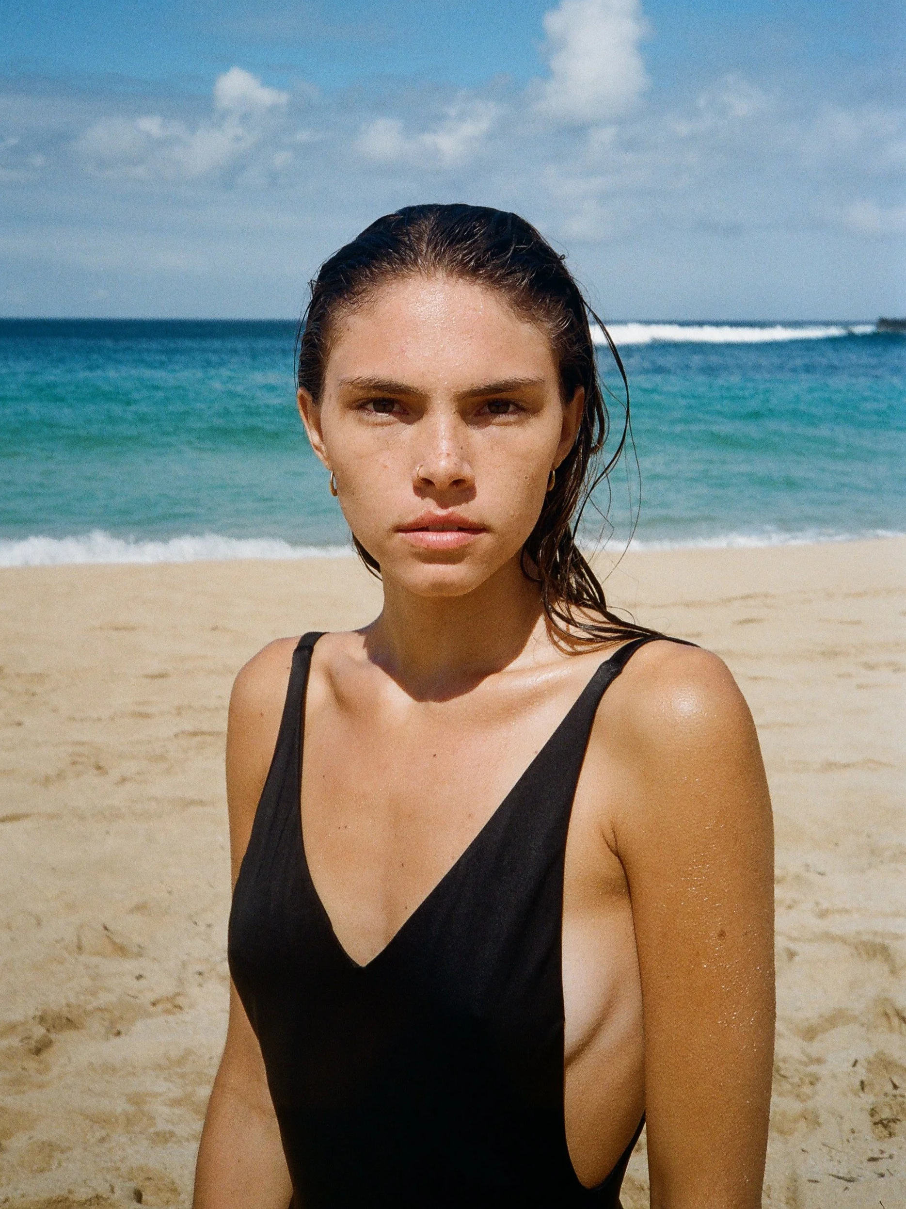 A woman with wet hair and tan skin wearing a black swimsuit, standing on a sandy beach with the ocean and blue sky in the background.