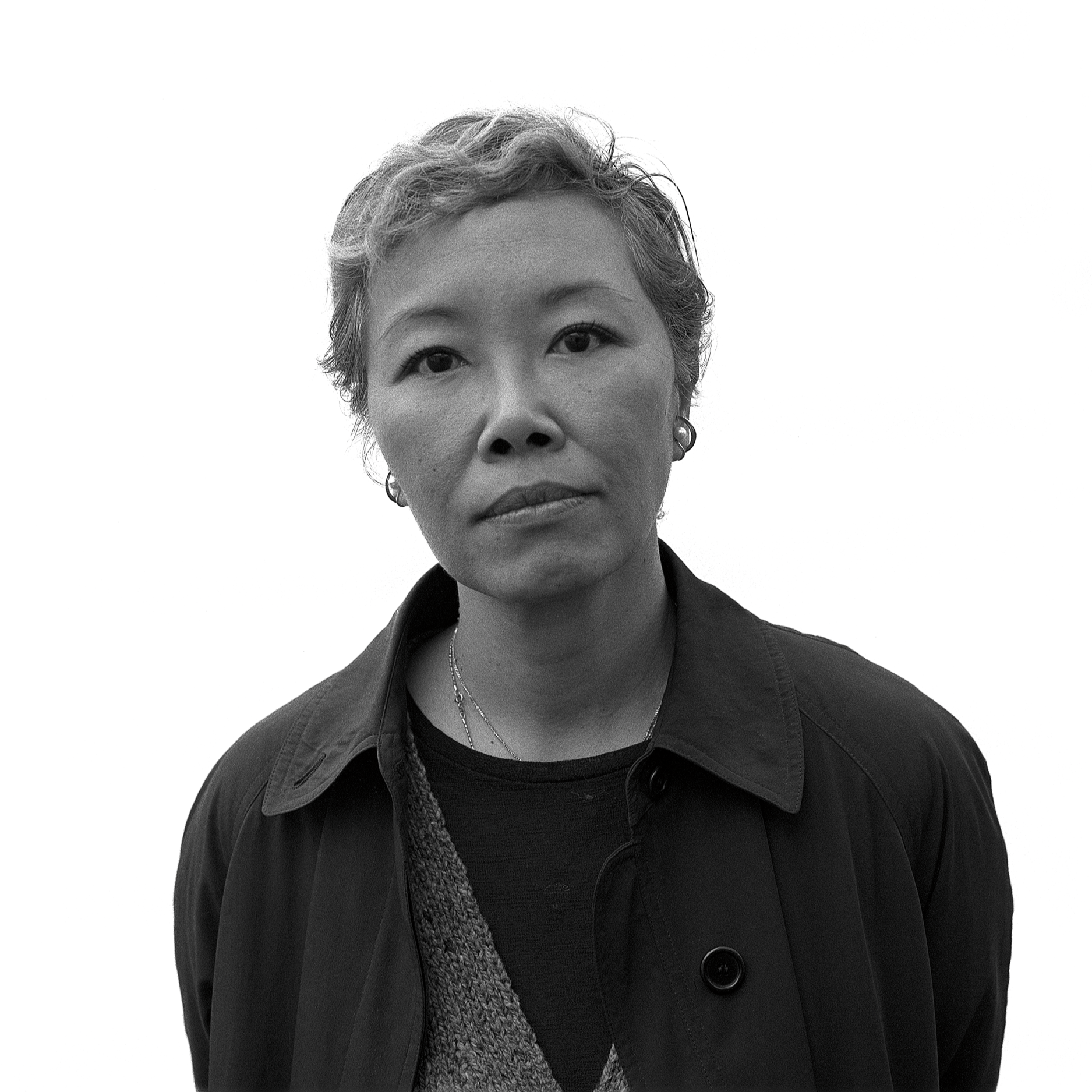 Black and white photo of a woman with short wavy hair, wearing a dark coat, a patterned scarf, earrings, and layered necklaces, against a plain background.