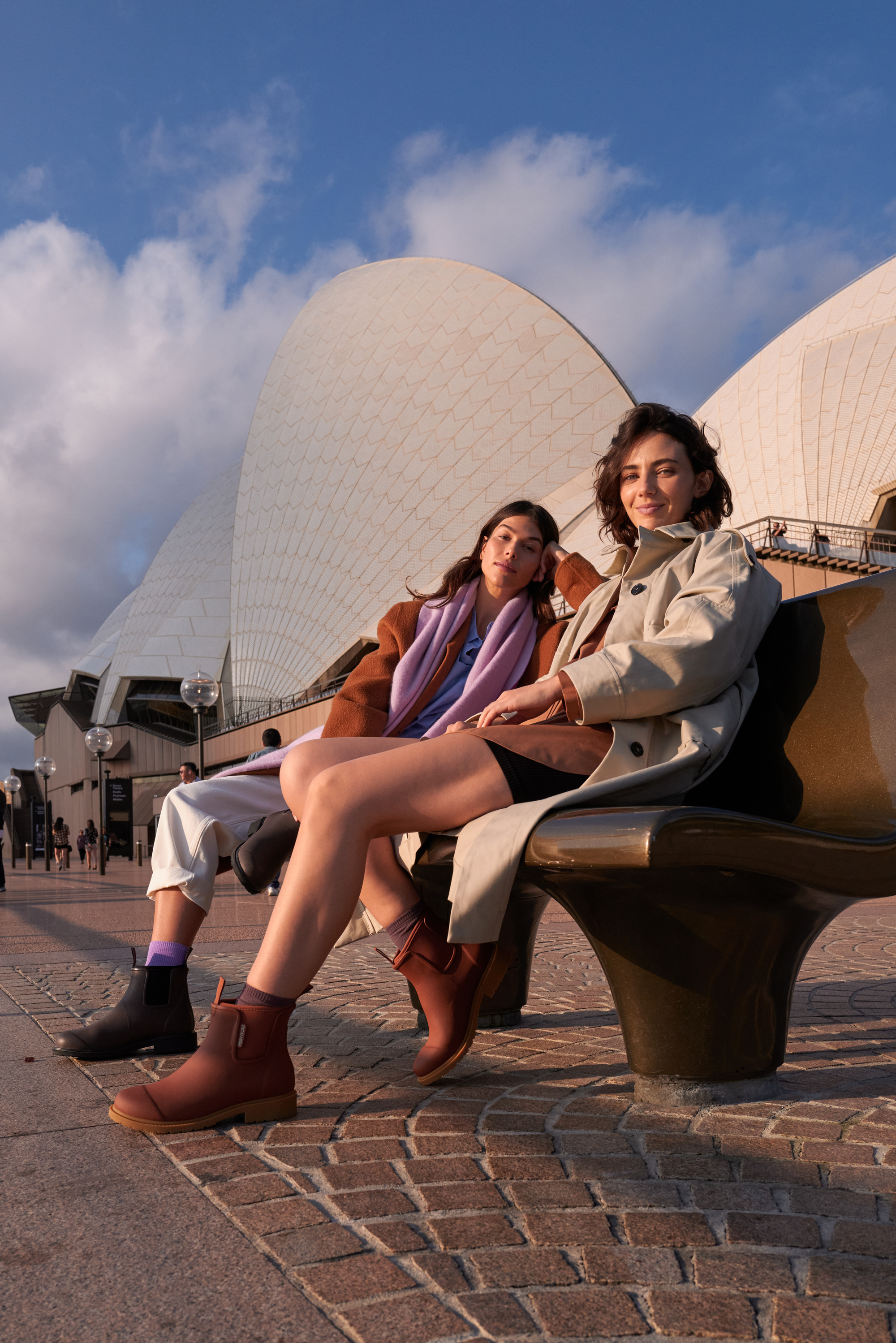 Two women sitting on a bench in front of the Sydney Opera House, with a few people walking in the background, during sunset.