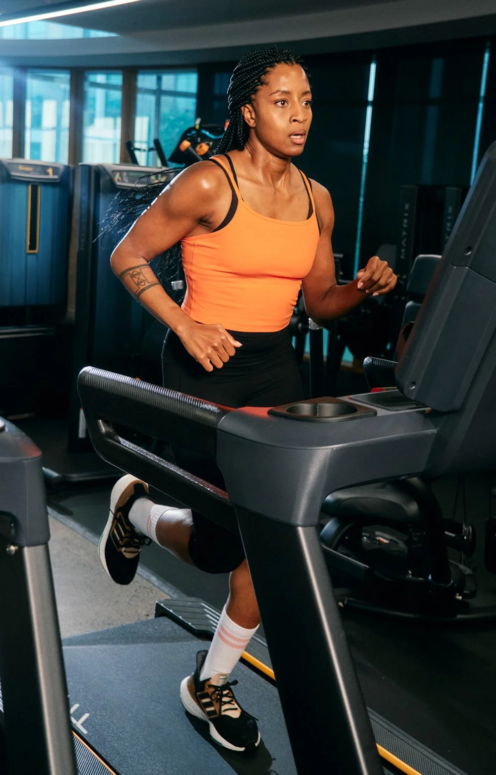 A woman running on a treadmill in a gym, wearing an orange tank top, black shorts, and running shoes.