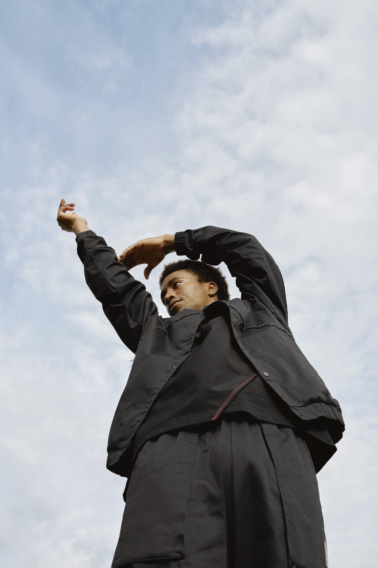 Person in black jacket with arms raised against cloudy sky.