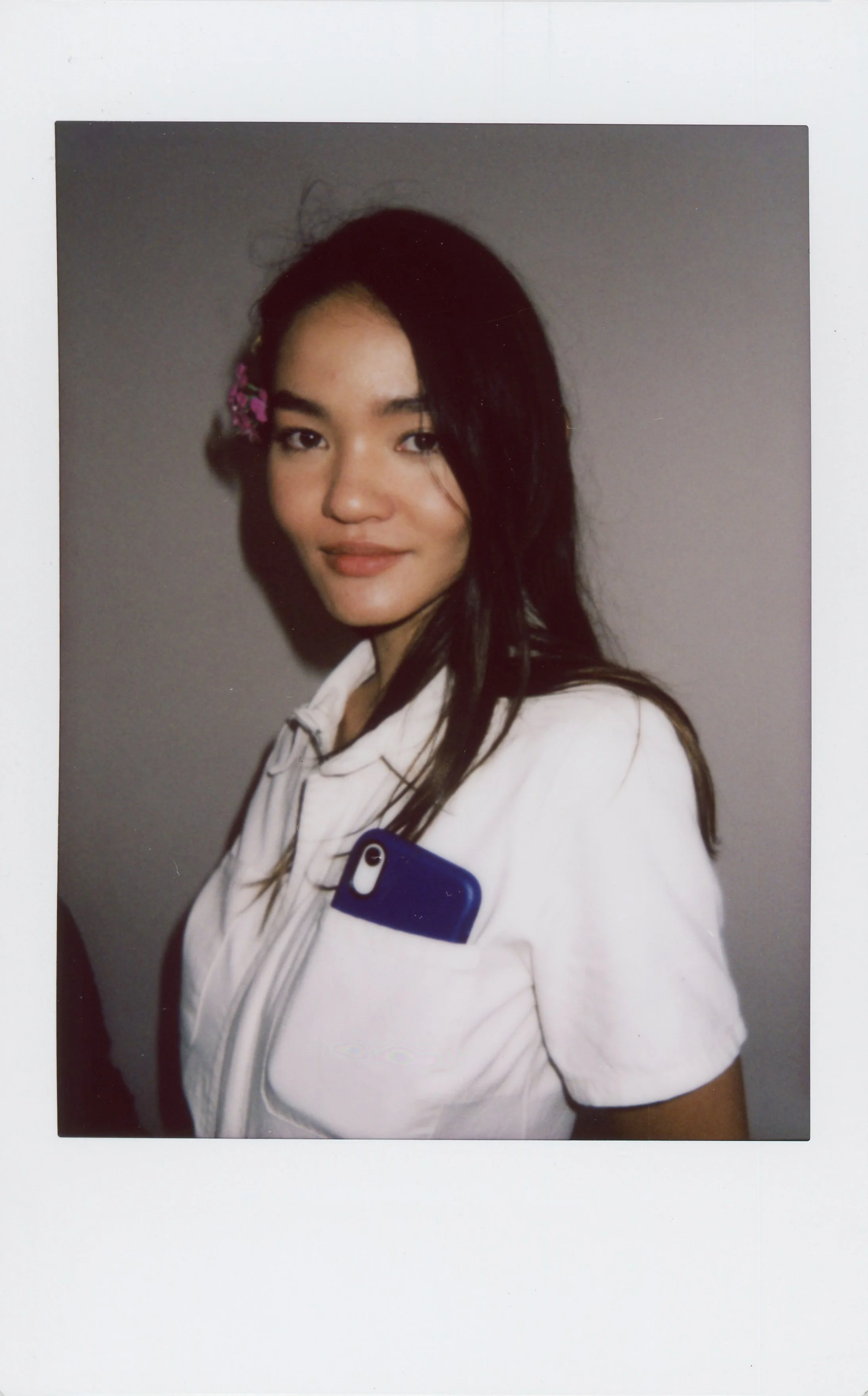 A young woman in a white jacket with a blue phone in her jacket pocket, standing against a plain gray background, looking at the camera and smiling