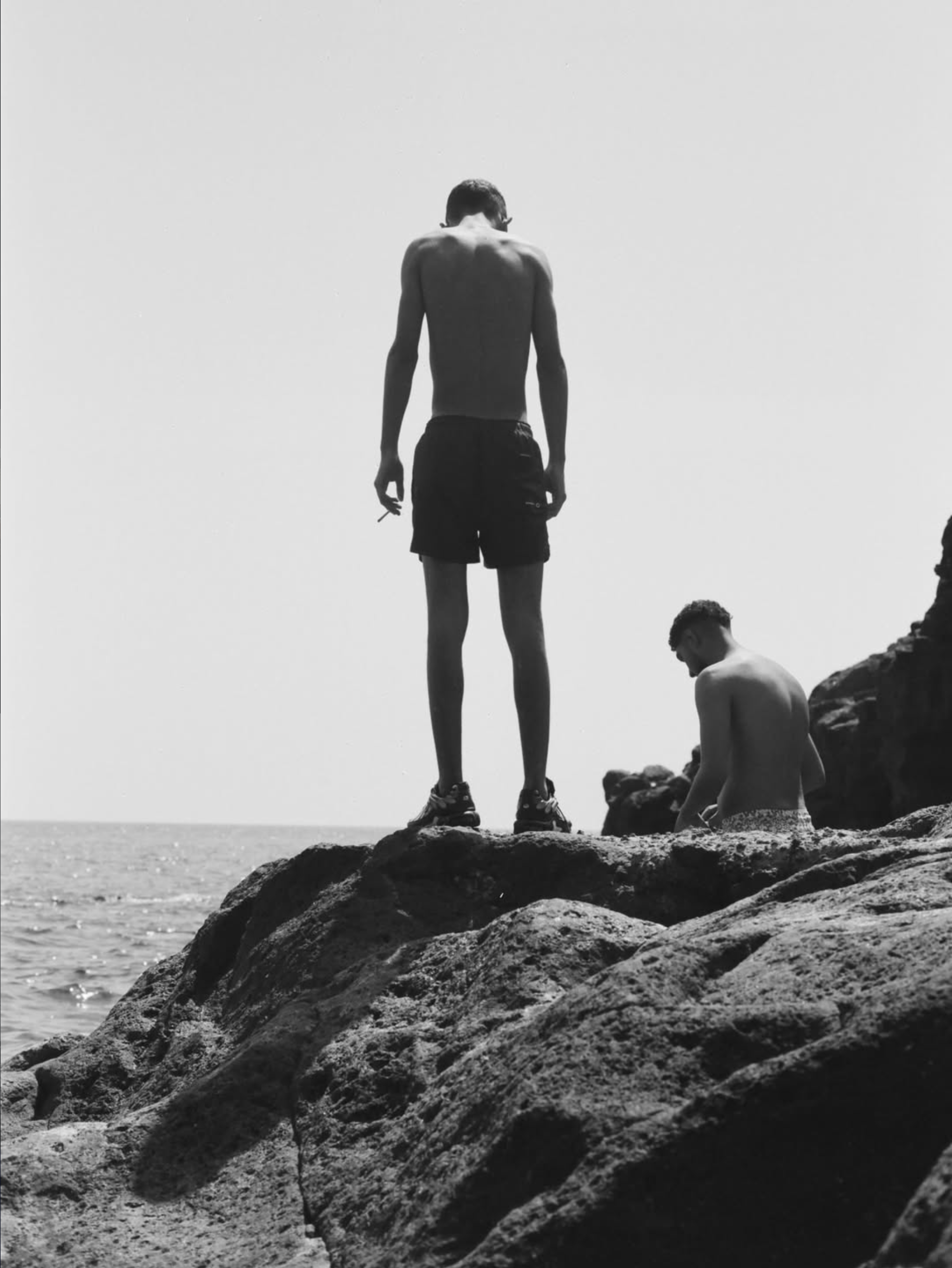 A black and white photo of a man standing on rocks at the beach, facing away from the camera, with two other people sitting nearby.