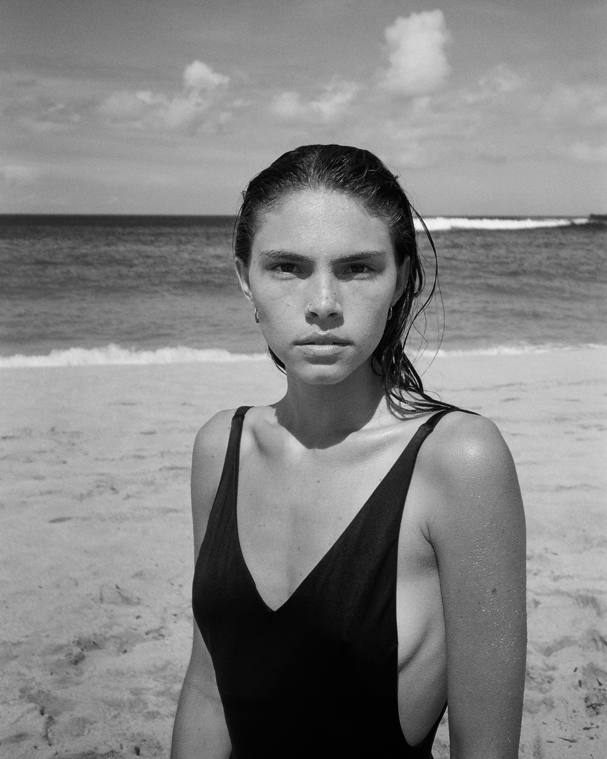Black and white photo of a woman in a swimsuit at the beach, with ocean waves and cloudy sky in the background.