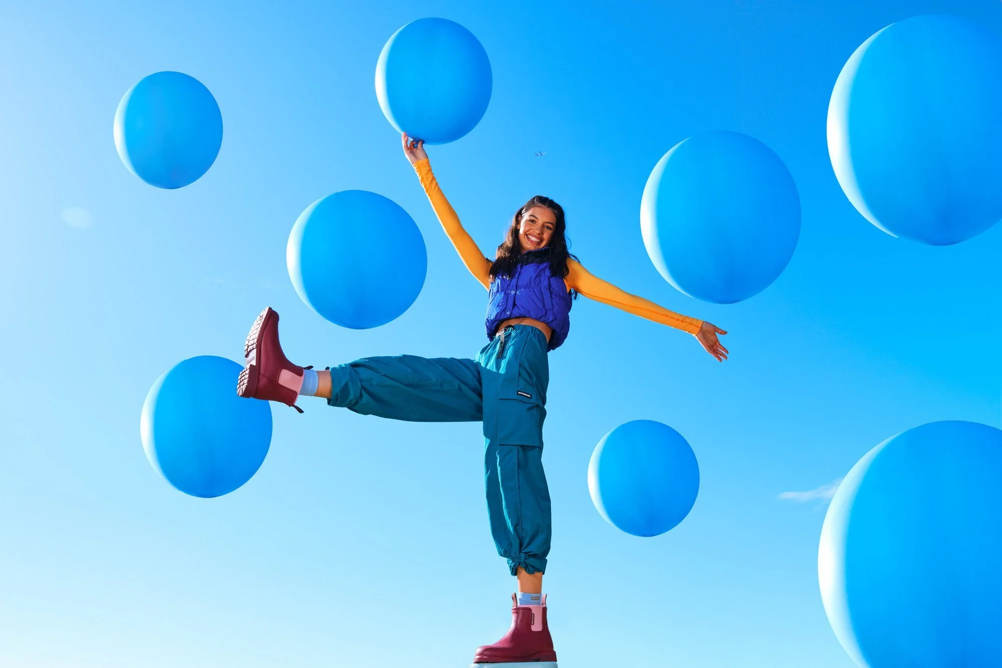 A woman in colorful outdoor clothing balancing on one leg while holding a blue balloon, surrounded by floating blue balloons in the sky.