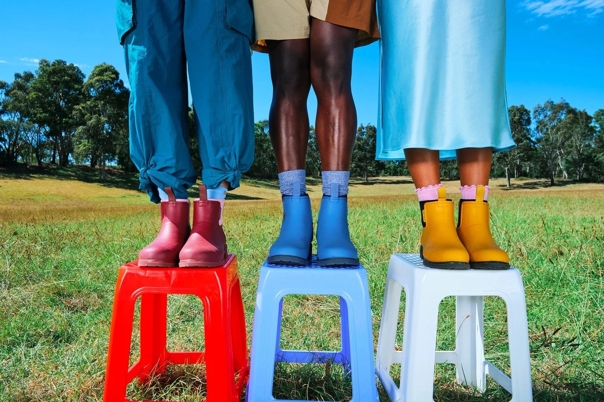 Close-up of three people standing on colorful stools outdoors, wearing rain boots, with trees and blue sky in the background.