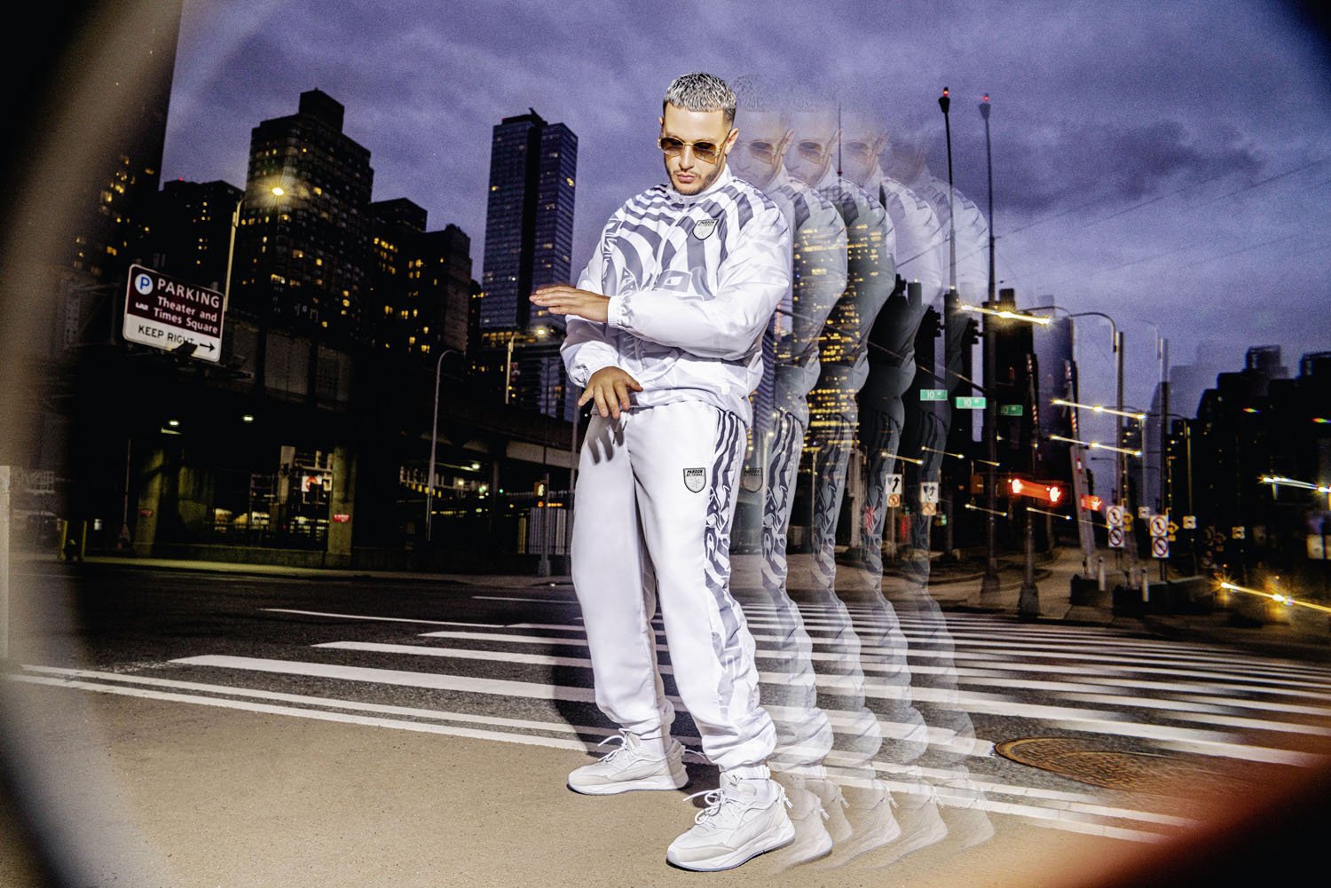DJ Snake with sunglasses and short hair, dressed in a shiny white tracksuit with patterns and logo, stands on a city street at dusk, with multiple exposures creating a ghosting effect, against a city skyline with tall buildings and streetlights.