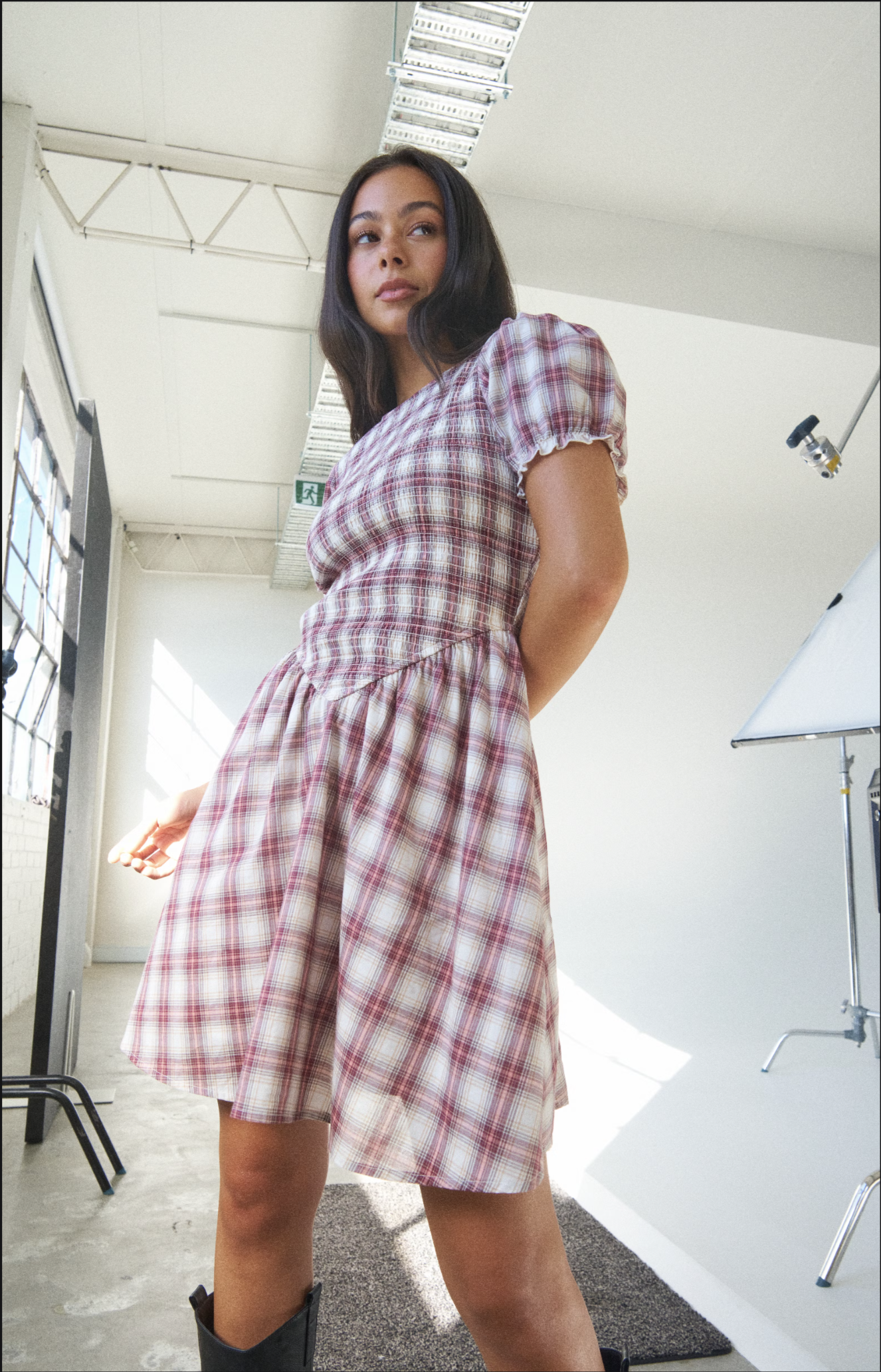 A woman in a pink and white plaid dress standing in a photography studio with a white backdrop and studio equipment visible.