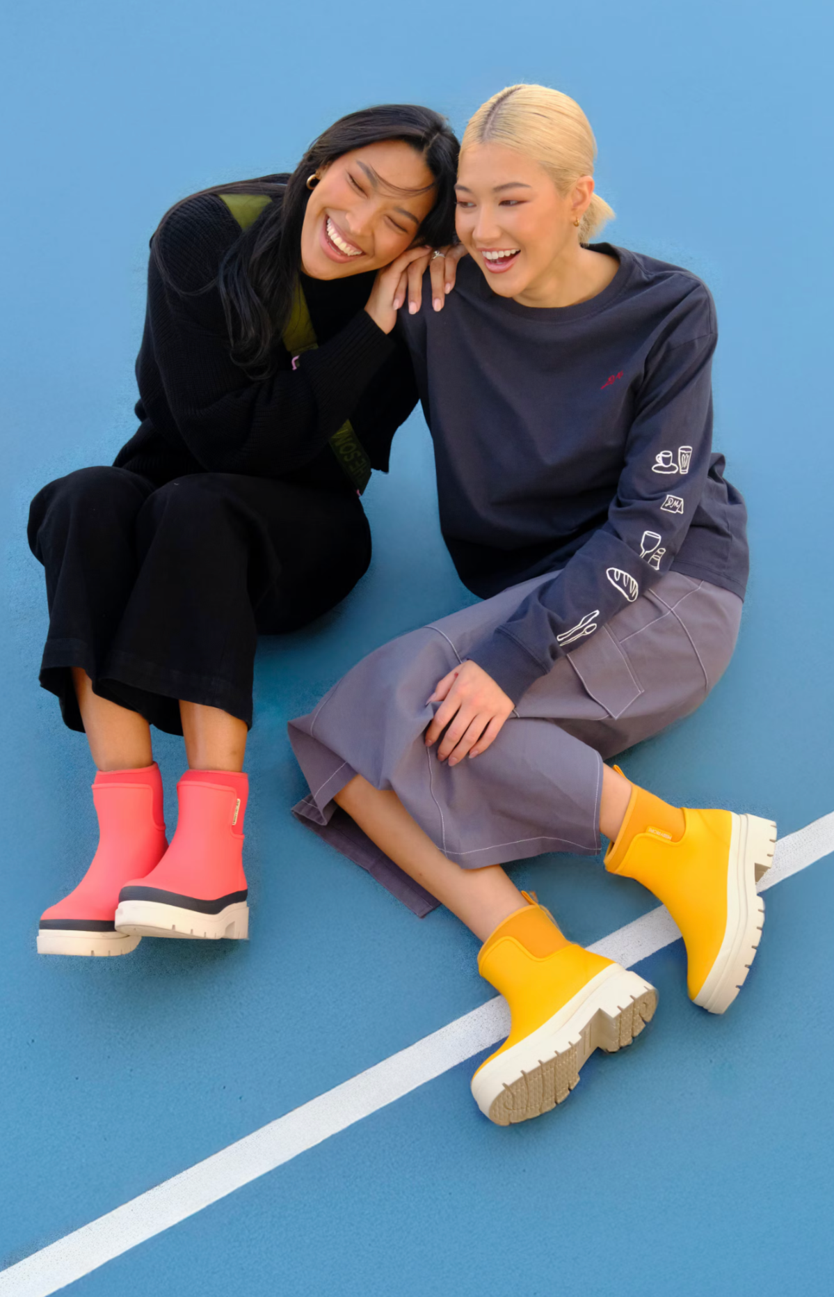 Two women sitting on a blue outdoor sports court, smiling and laughing together, wearing colorful rain boots and casual clothing.