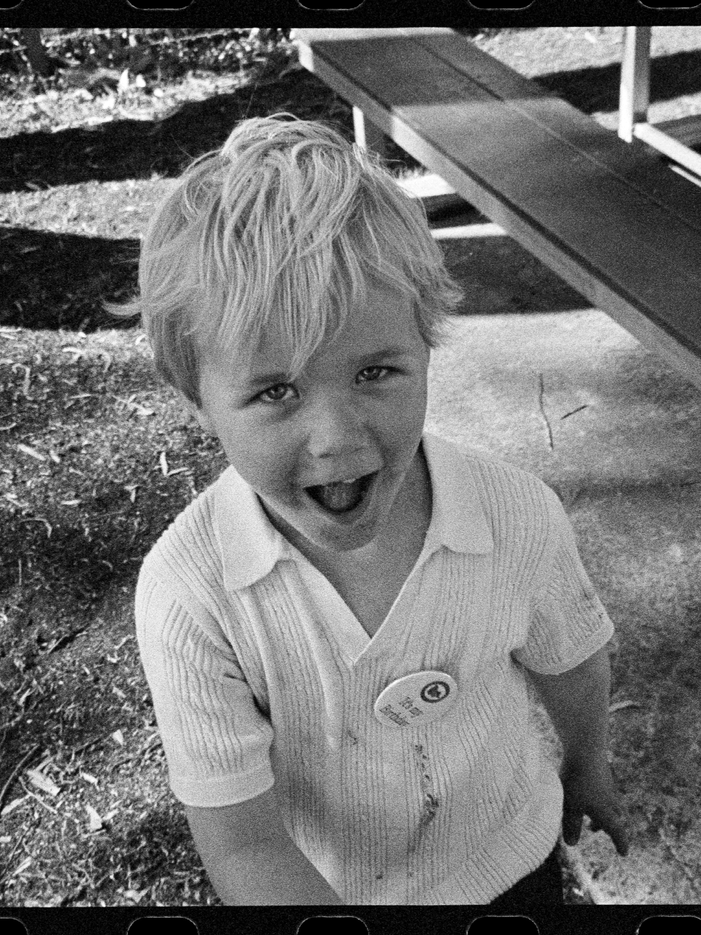 A young boy with light-colored, messy hair, wearing a collared shirt, smiling with his mouth open, standing outdoors near a picnic table.