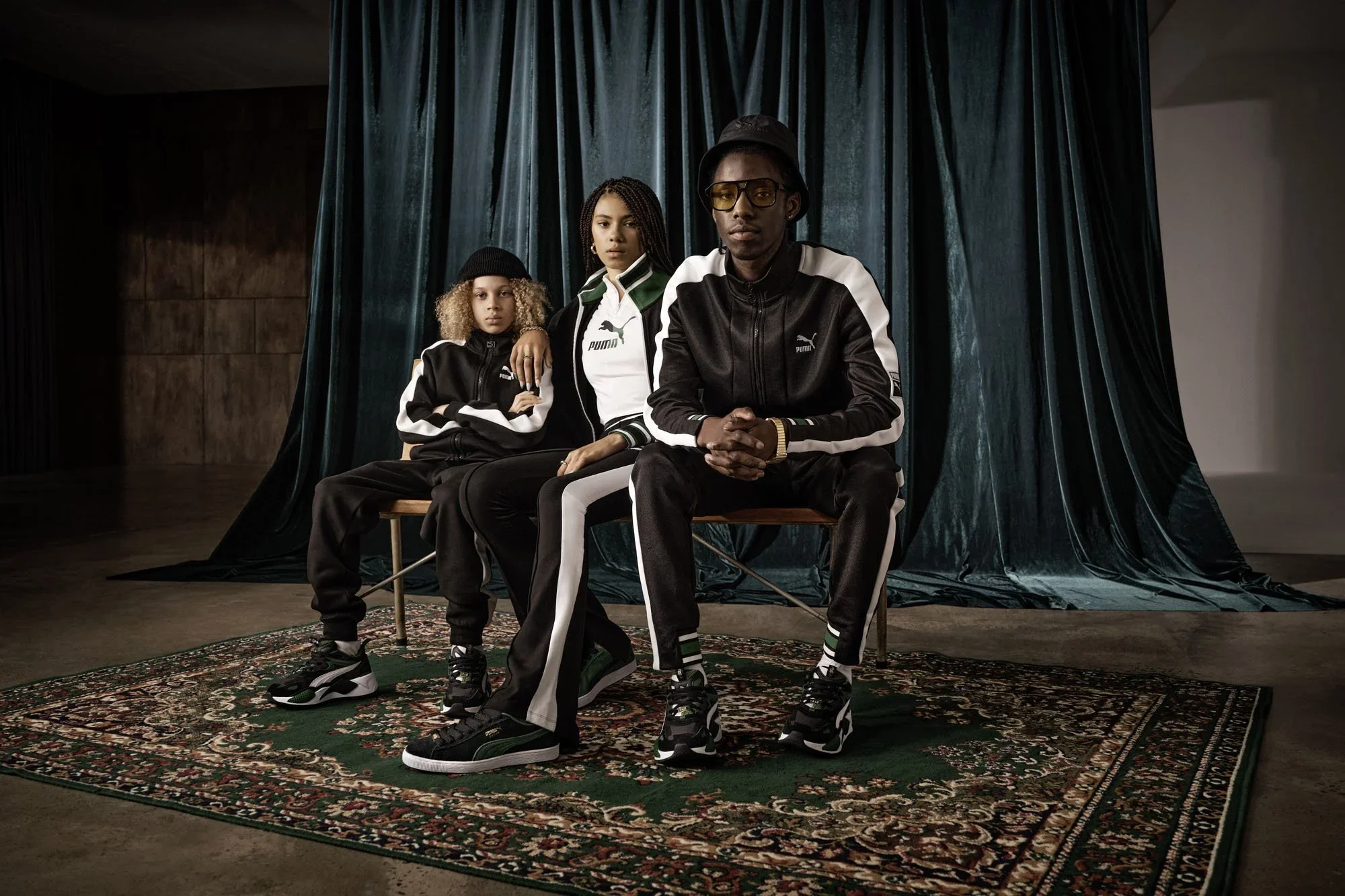 Group of four young people sitting on chairs in front of a teal curtain, wearing Puma athletic wear, sneakers, and casual accessories, in a studio setting with a patterned rug and wooden walls.