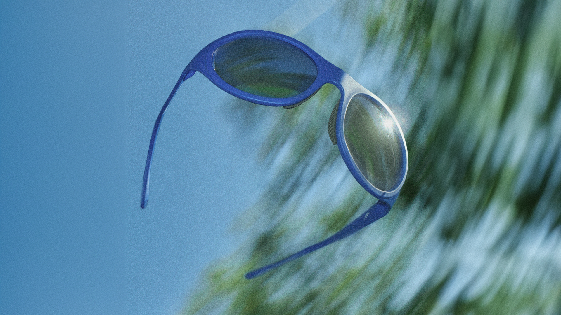 Blue sunglasses with dark lenses on a reflective surface, with a blurred background of green palm leaves.