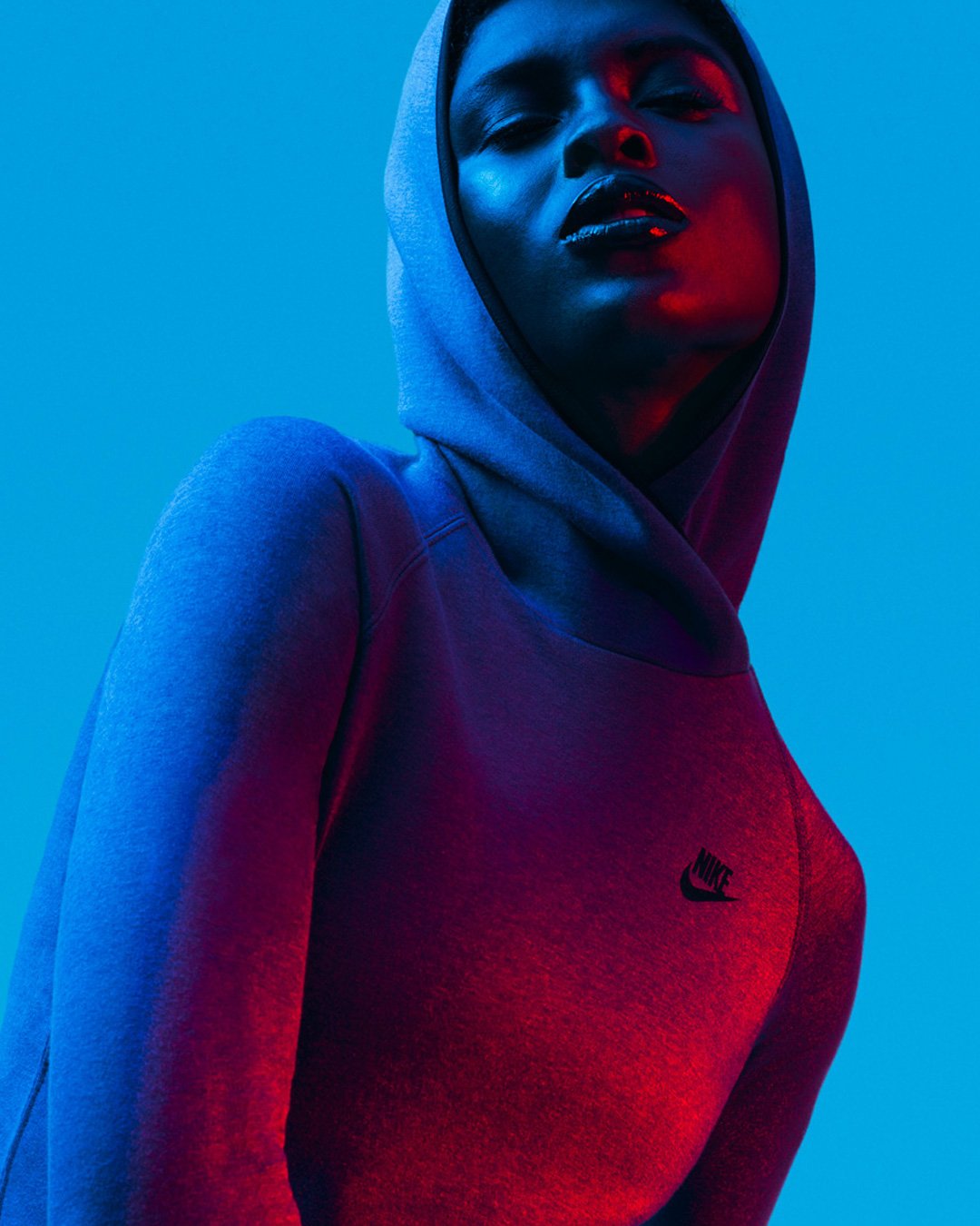 A woman wearing a gray Nike hoodie poses against a blue background with dramatic red and blue lighting highlighting her face and hoodie.