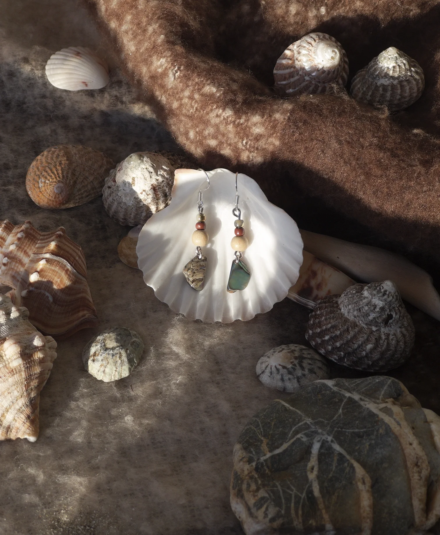 Shells and earrings placed on a brown fabric surface surrounded by various seashells and stones.