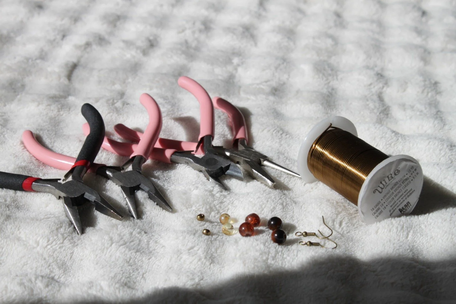 A spool of gold thread, three pairs of jewelry pliers with pink and black handles, several small beads in different colors, two small hooks with beads, and two small metal components on a white textured surface.