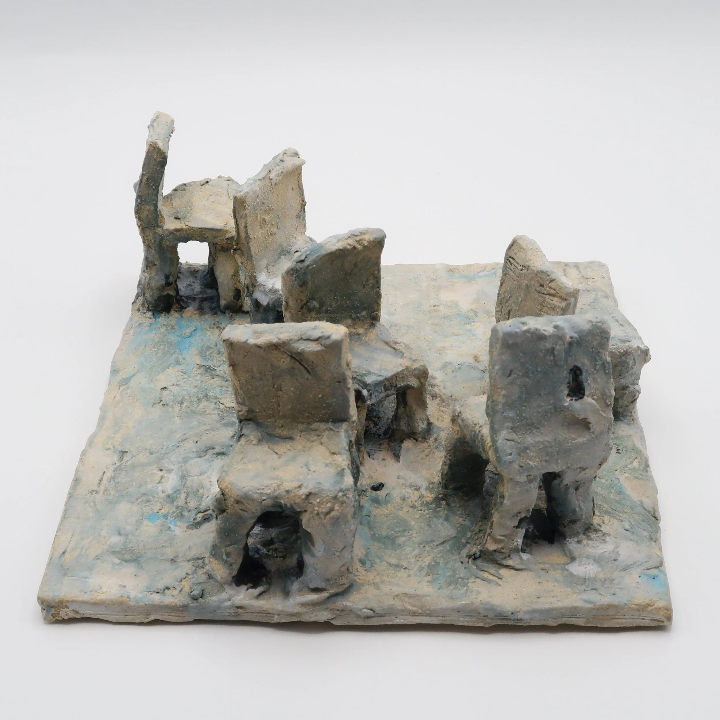 Close-up of a small, weathered sculpture made of clay or similar material, depicting an abstracted horse or animal figure, mounted on a rectangular base against a plain white background.