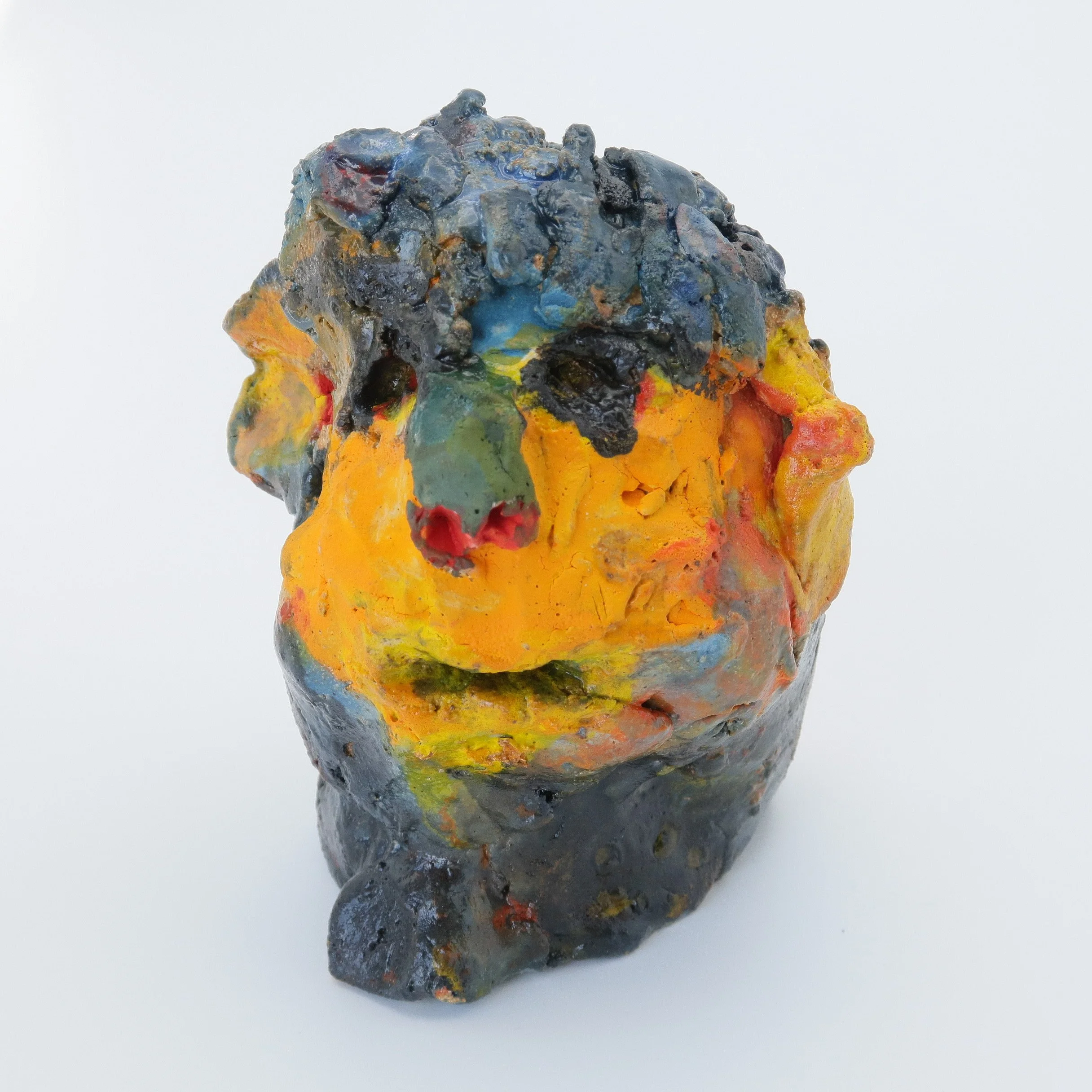 A colorful, textured sculpture made of clay, painted with various colors including blue, yellow, orange, and black, with an irregular shape and rough surface.