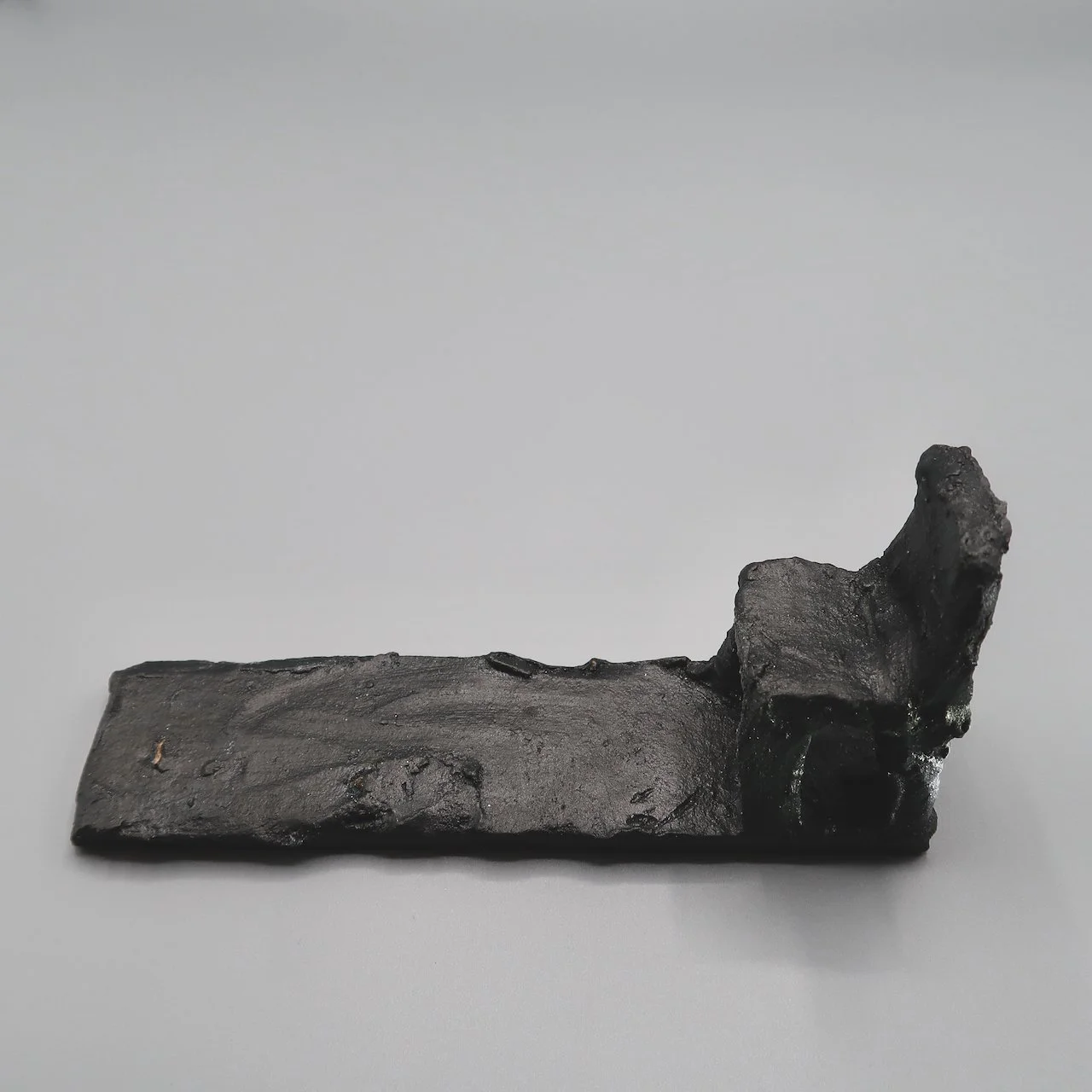 A small, black, textured object made of a rough material, possibly metal or ceramic, resting on a plain light gray surface.