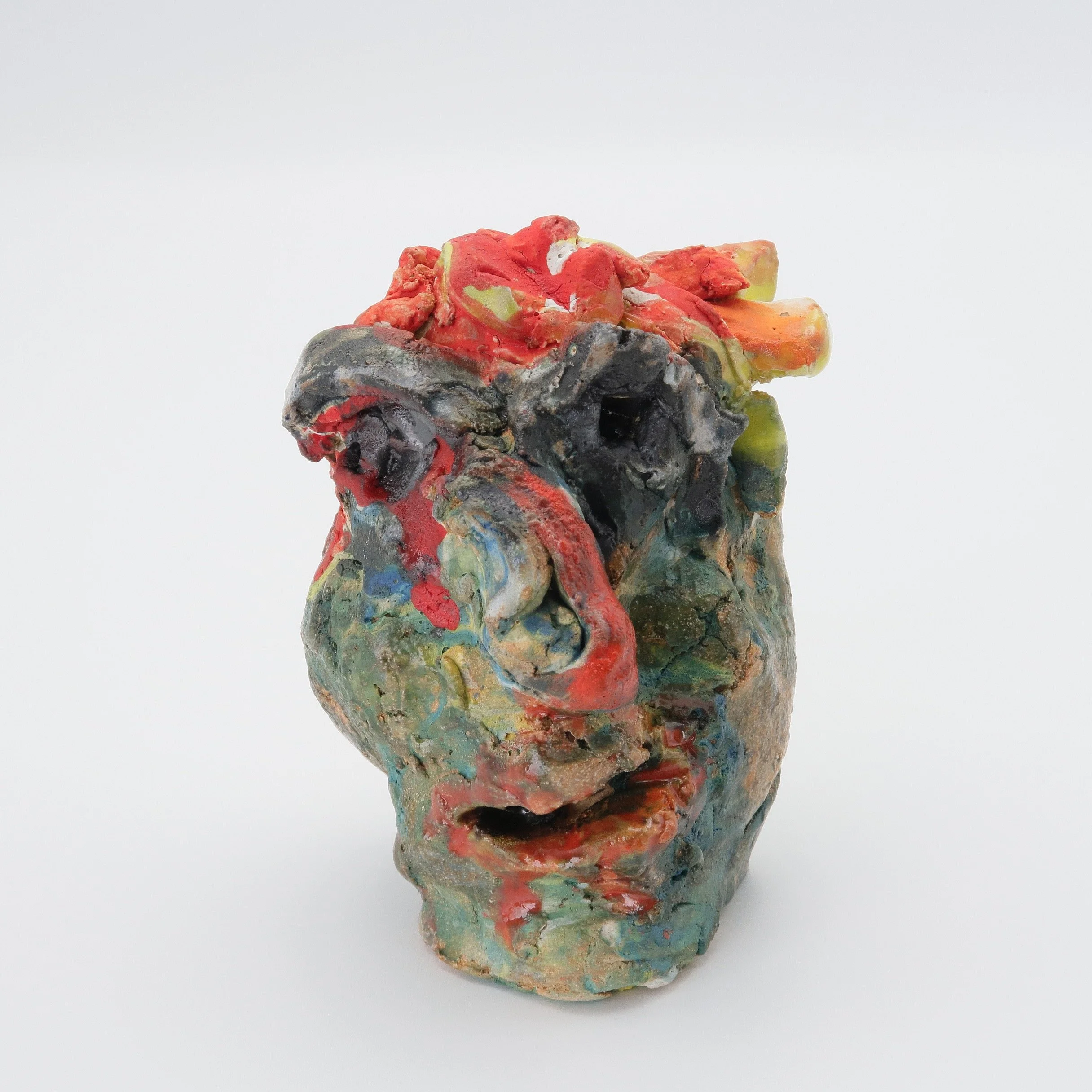 A colorful, abstract sculpture made of clay or similar material, featuring vibrant red, yellow, green, orange, and black colors with a textured and layered appearance, set against a plain white background.