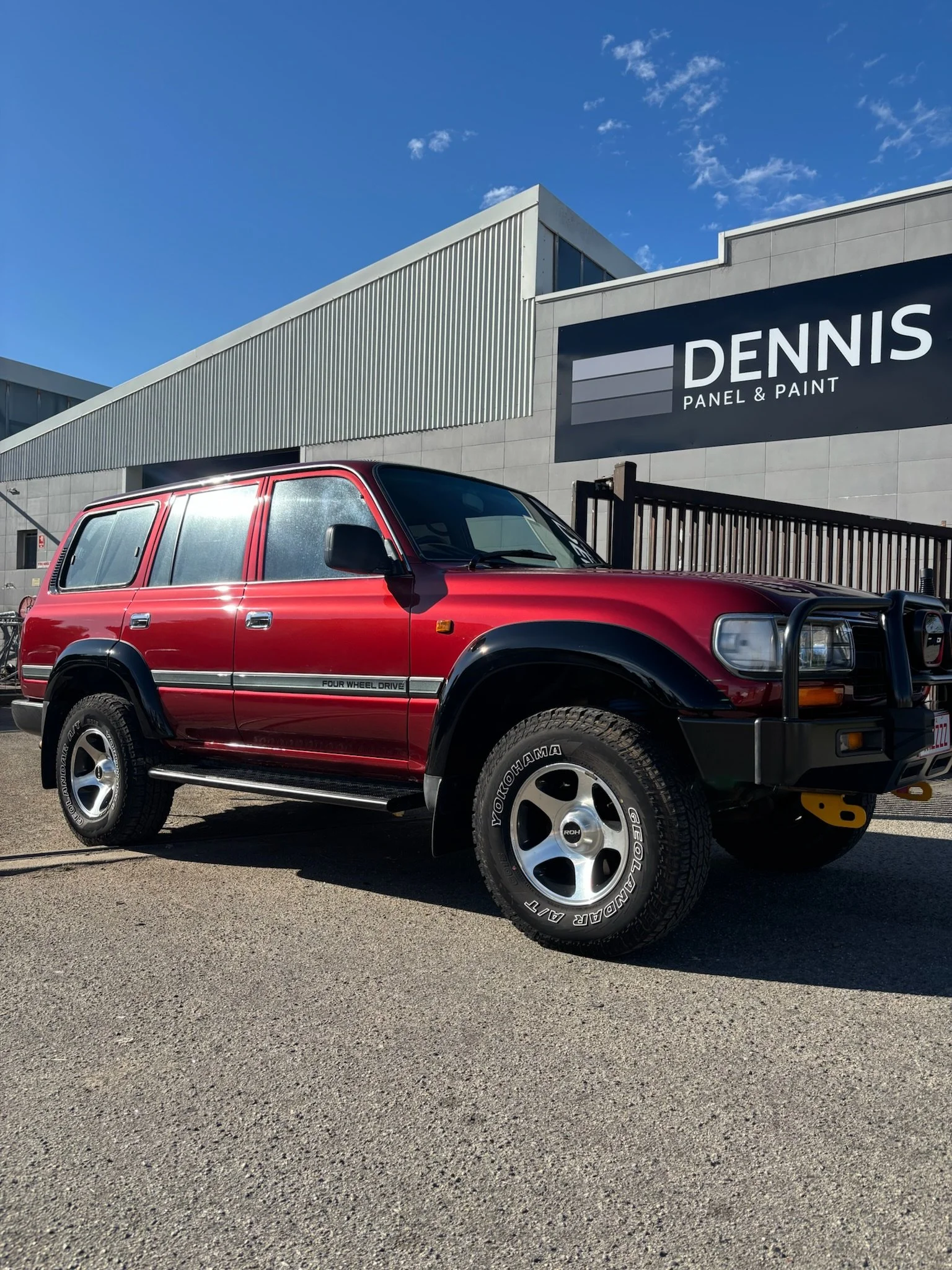 Red four-wheel-drive vehicle parked outside a building with a sign reading "Dennis Panel & Paint" against a blue sky with a few clouds.