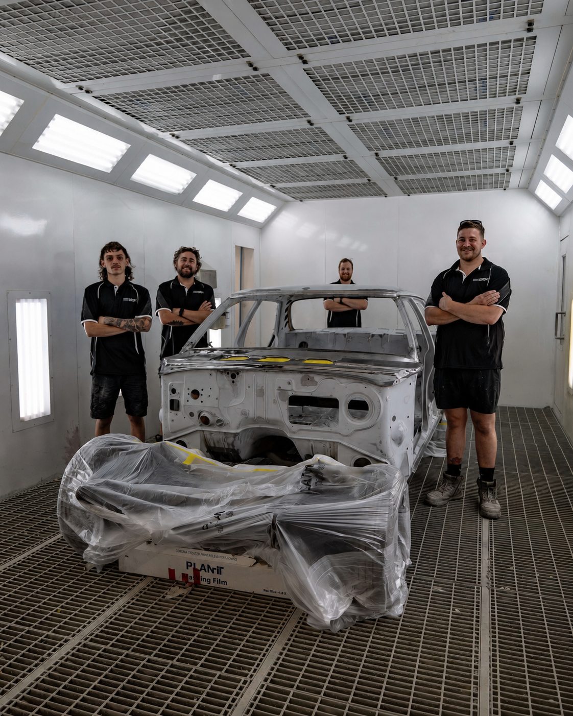 Four people standing in a paint booth around an unfinished car body and parts, with equipment and lighting overhead.