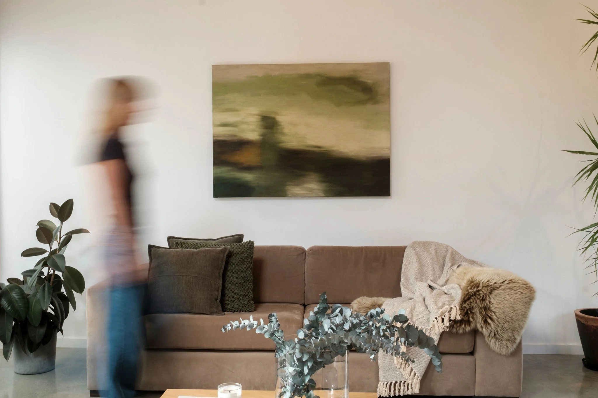 Living room with beige sofa, abstract painting, green plants, glass vase with eucalyptus, beige and furry blanket, and a person walking by.