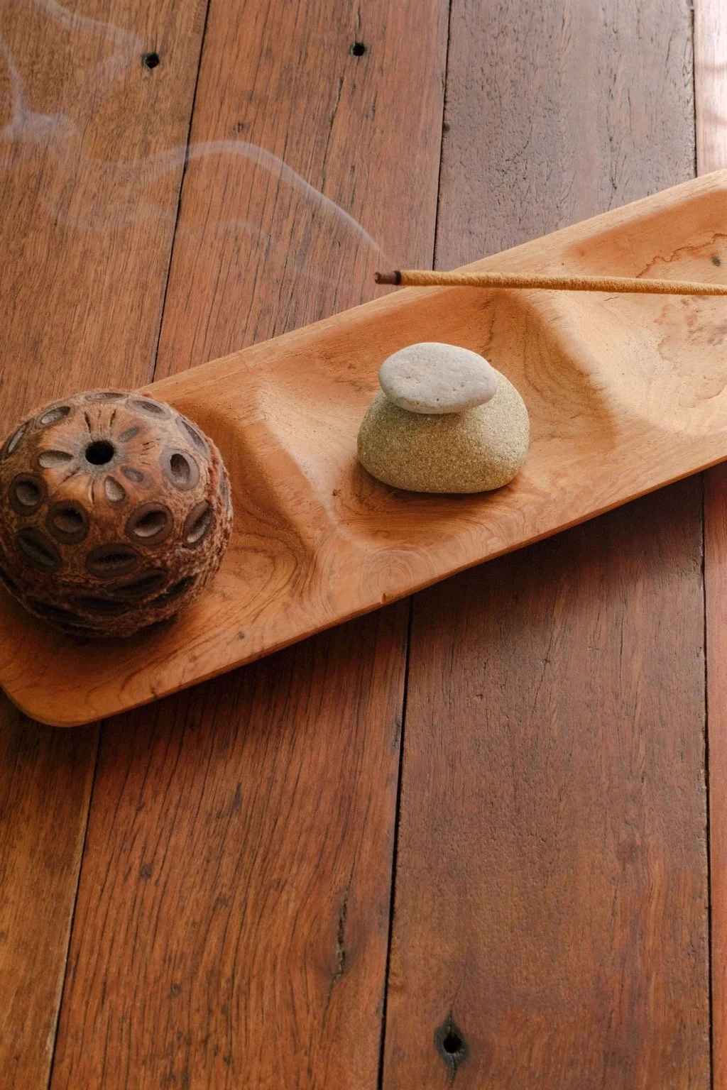 A wooden tray on a wooden floor contains a dried round seedpod, two stacked stones, and a wooden incense stick emitting smoke.