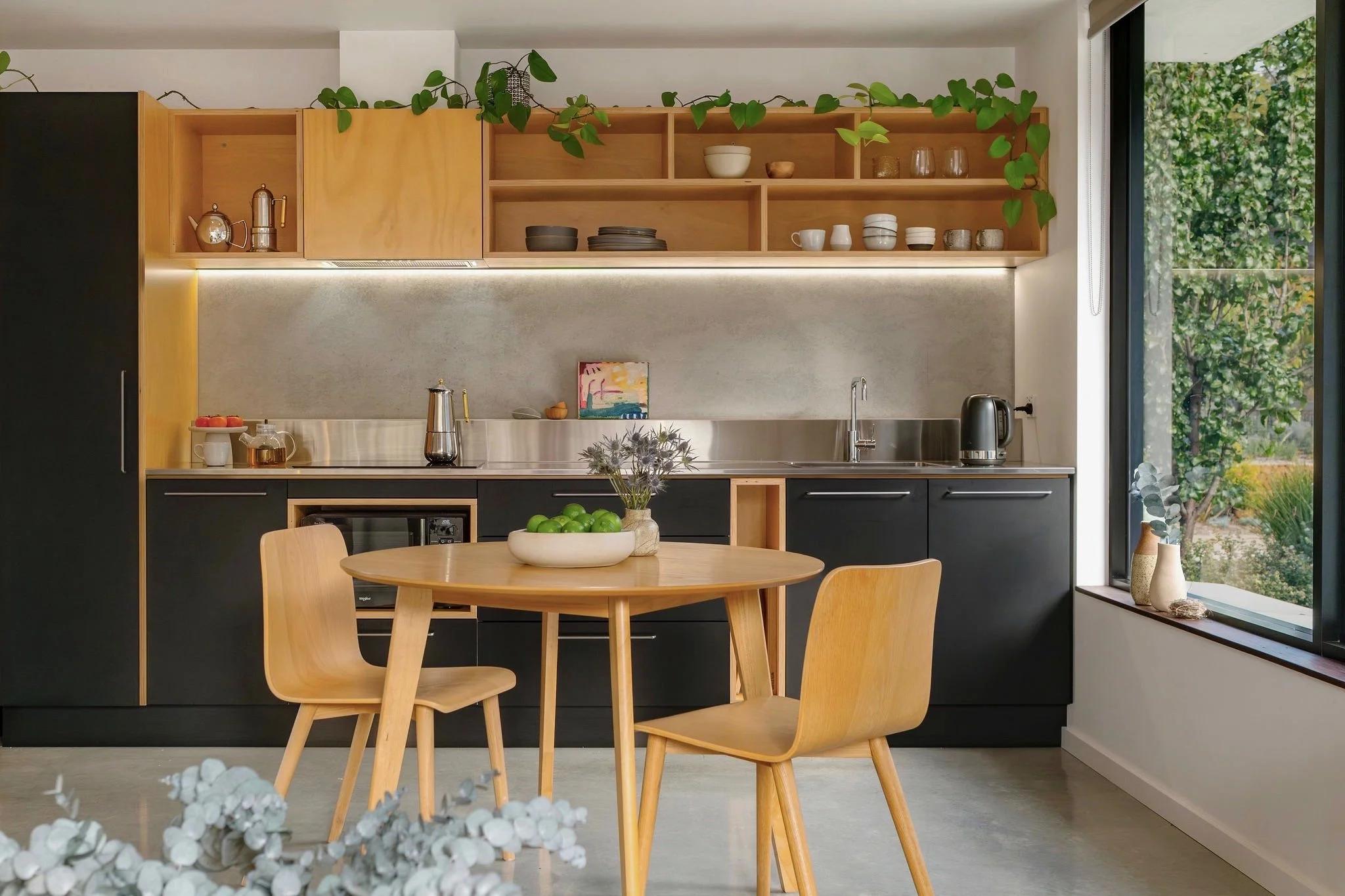 Modern kitchen with open wooden shelves displaying dishes, cups, and glassware, a round dining table with wooden chairs, large window showing outdoor greenery, and a neutral color palette.
