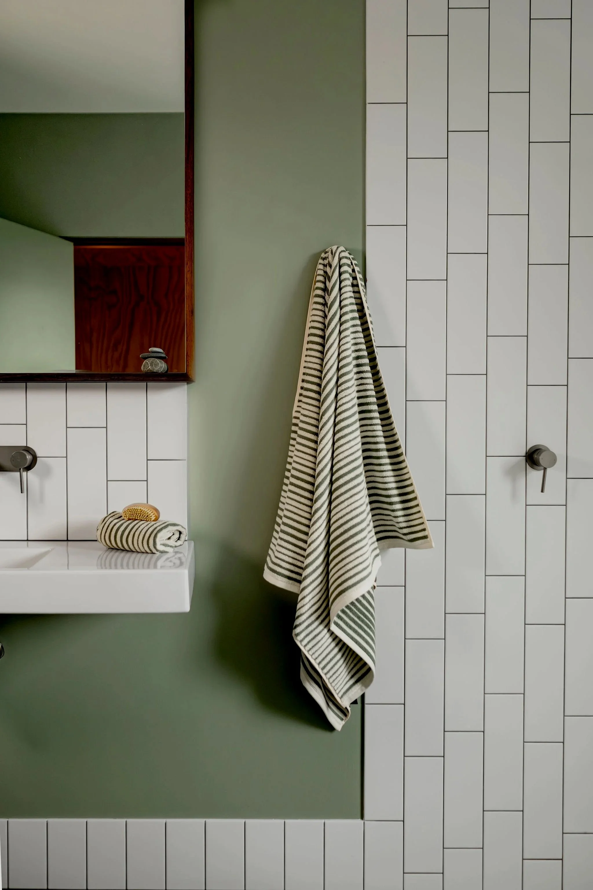 A bathroom with a green wall, a rectangular mirror, white tile walls, a striped towel hanging, and a stacked set of towels on a white sink.