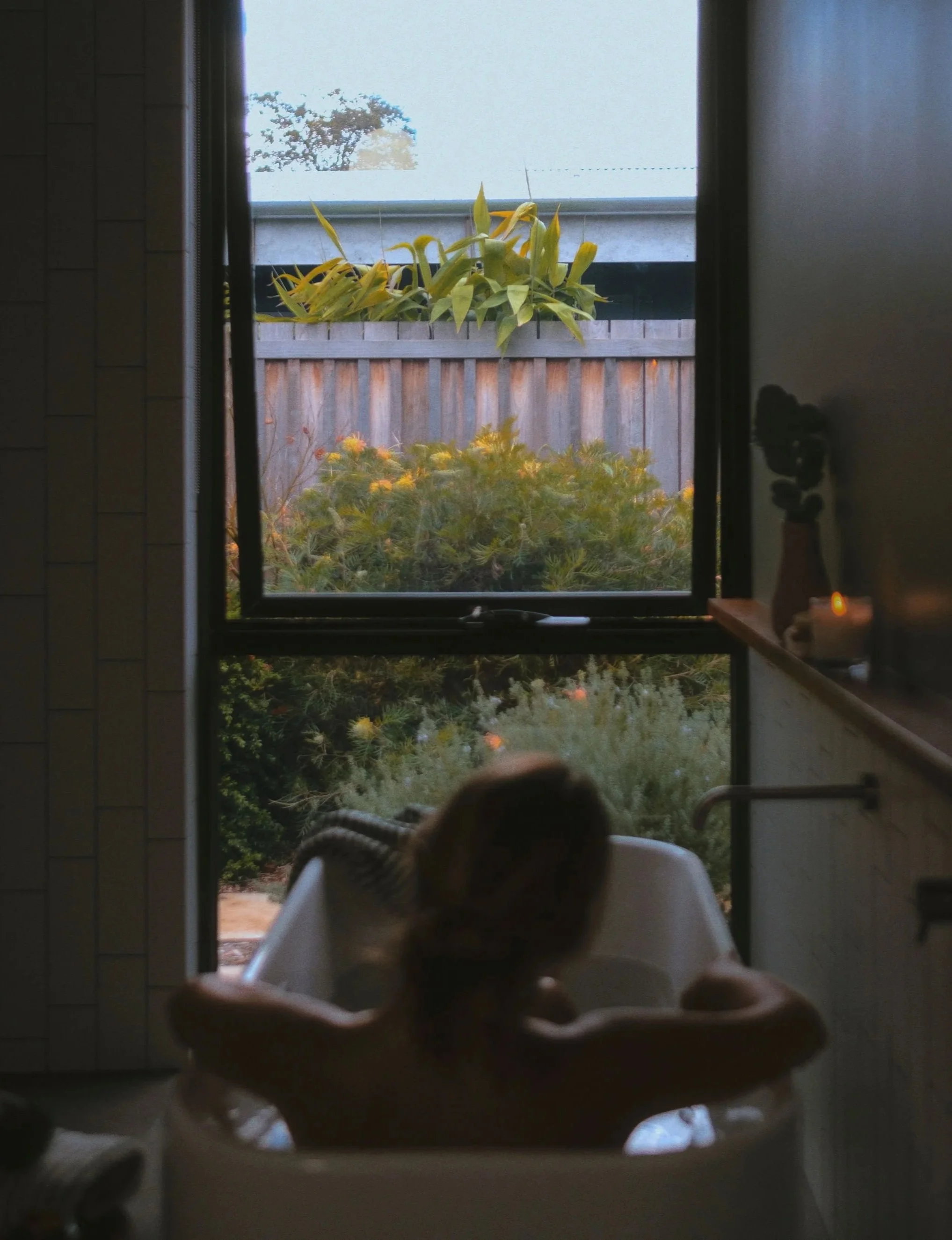 Child sitting in a high chair looking out a window at a garden with plants and a wooden fence.