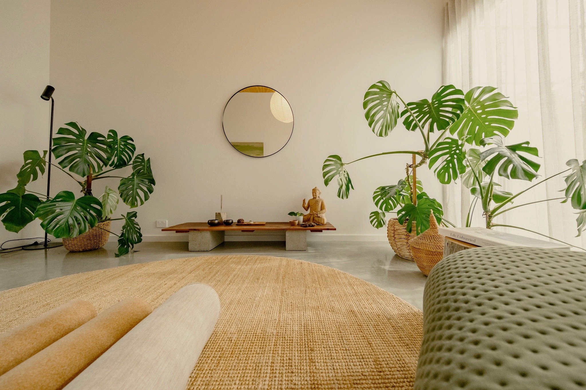 Living room with large Monstera plants, a wooden table with a Buddha statue, decorative bowls, and a mirror on the wall, with natural light coming through sheer curtains.