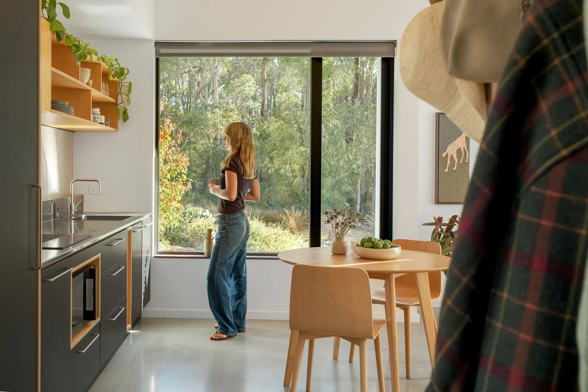 A woman with long blonde hair wearing a black t-shirt and loose jeans standing in a kitchen near a large window, holding a mug. The kitchen has wooden shelves with dishes on the left, and a round wooden dining table with a bowl of green apples and a vase with dried flowers. Part of a person wearing a large straw hat and plaid shirt is visible in the foreground, and outside the window, there is greenery with trees.