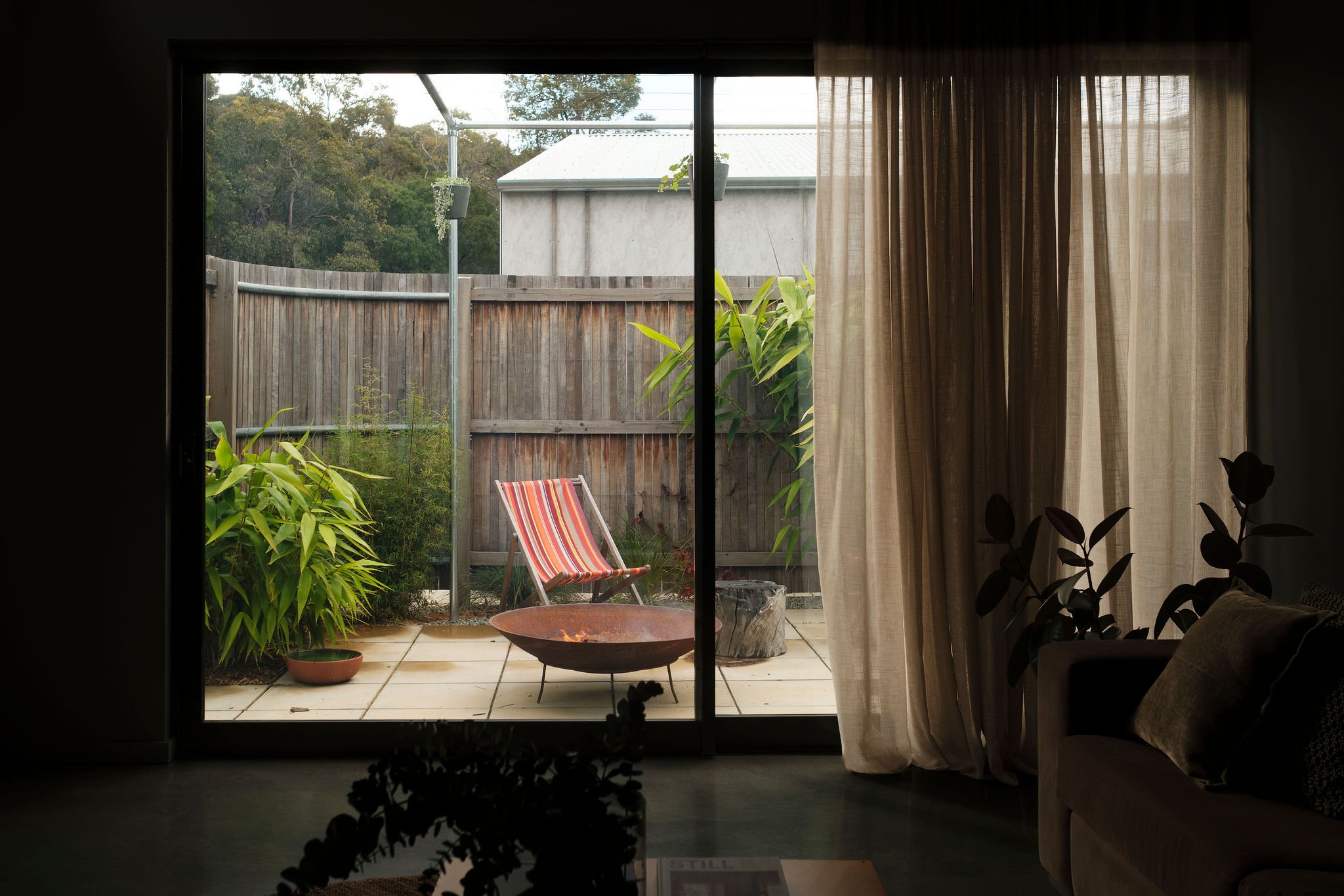 View from inside a room looking out through a sliding glass door to a small backyard patio with a colorful striped chair, potted plants, and a wooden fence, with trees in the background.