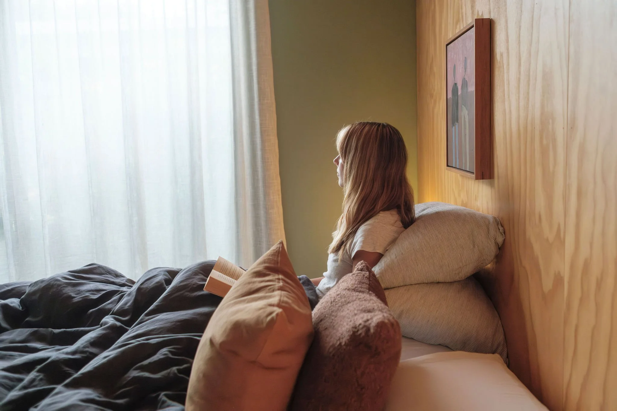 A woman with blondechair sitting on a bed, reading a book, in a bedroom with a plywod wall and a window with sheer curtains
